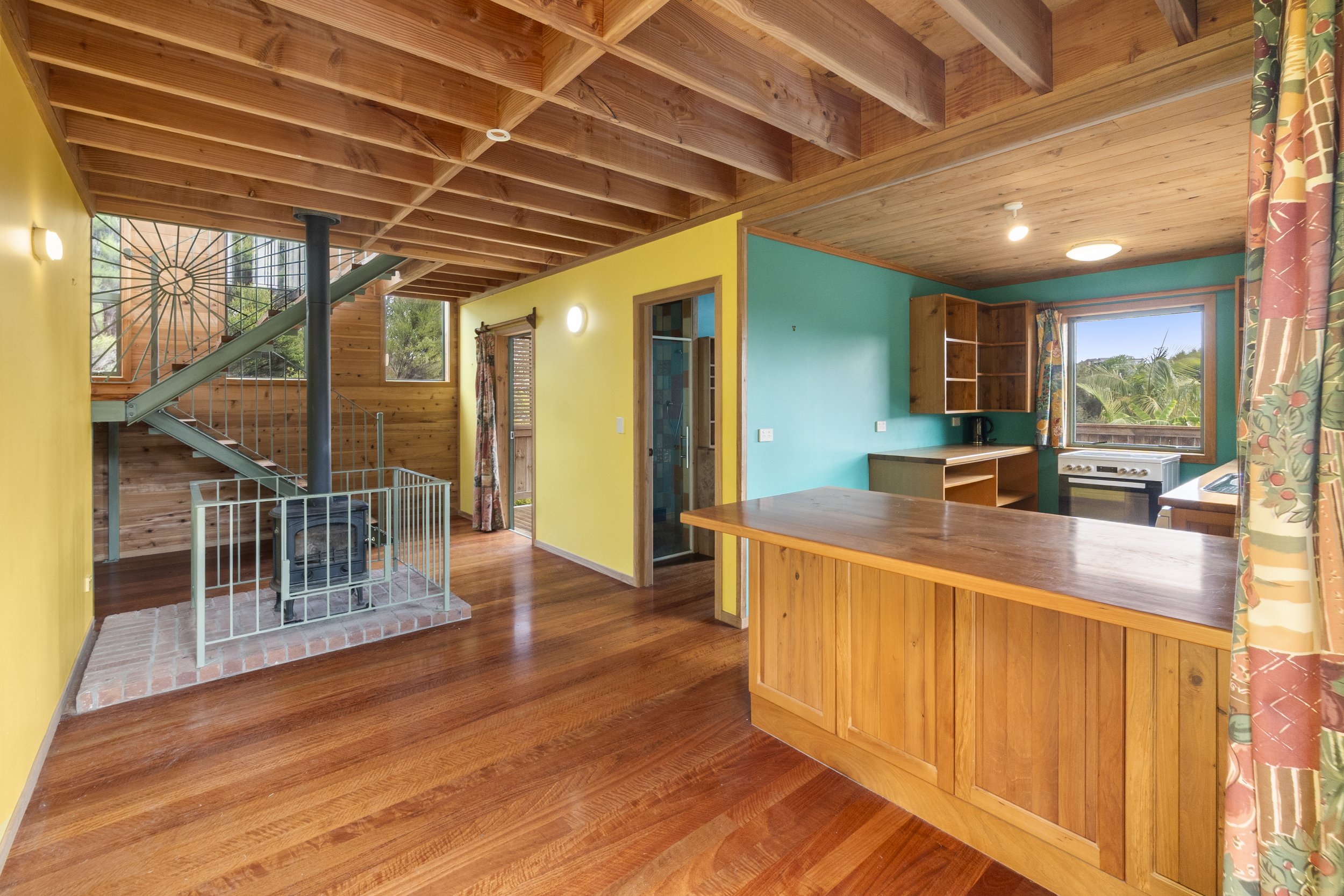 Interior of a wooden home featuring a cozy room with a wood-burning stove, visible staircase, open kitchen with wooden cabinets, and vibrant yellow and teal walls. The space has exposed ceiling beams and hardwood flooring.