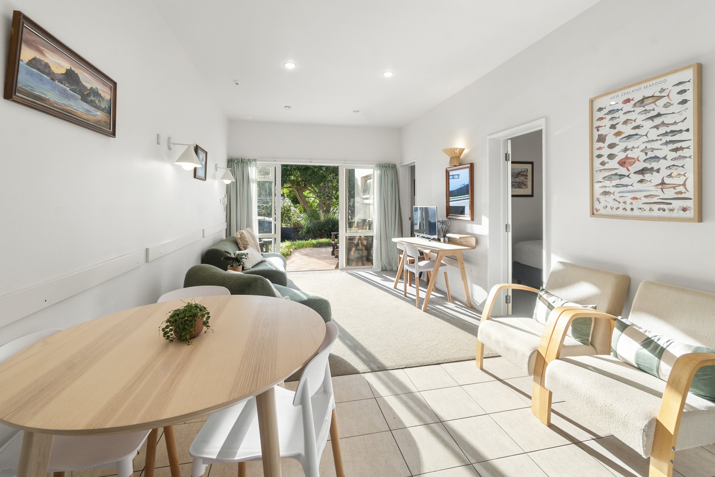 Modern living room with a dining table, sofa, desk, and chairs, featuring large windows and doors leading to a garden. Walls have art and a seafood poster. Bright and airy decor.