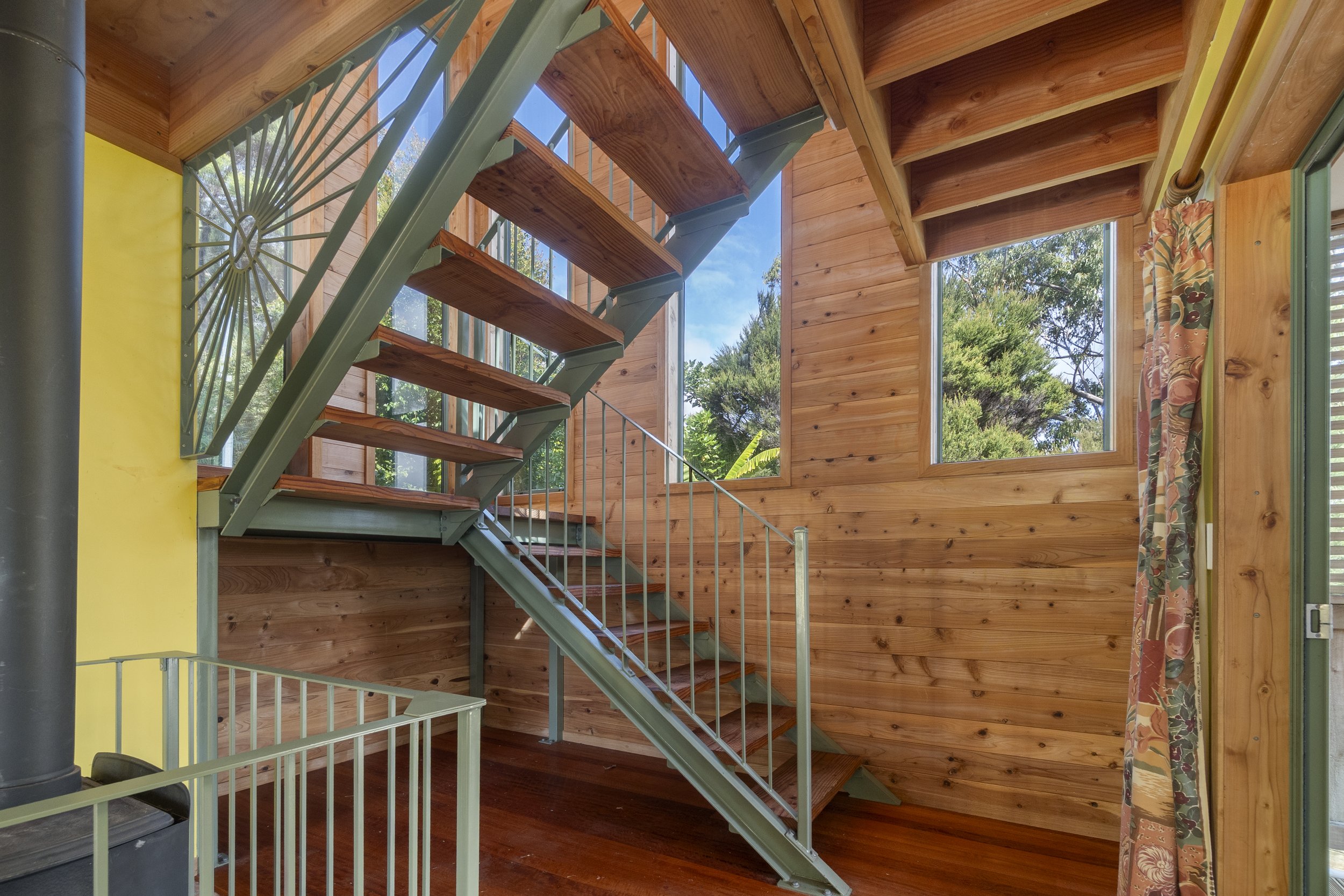 Interior view of a wooden staircase with metal railings, large windows, and a wooden-paneled wall. Sunlight filters through, highlighting the natural wood and greenery visible outside. A metal stove is partially visible on the left side.