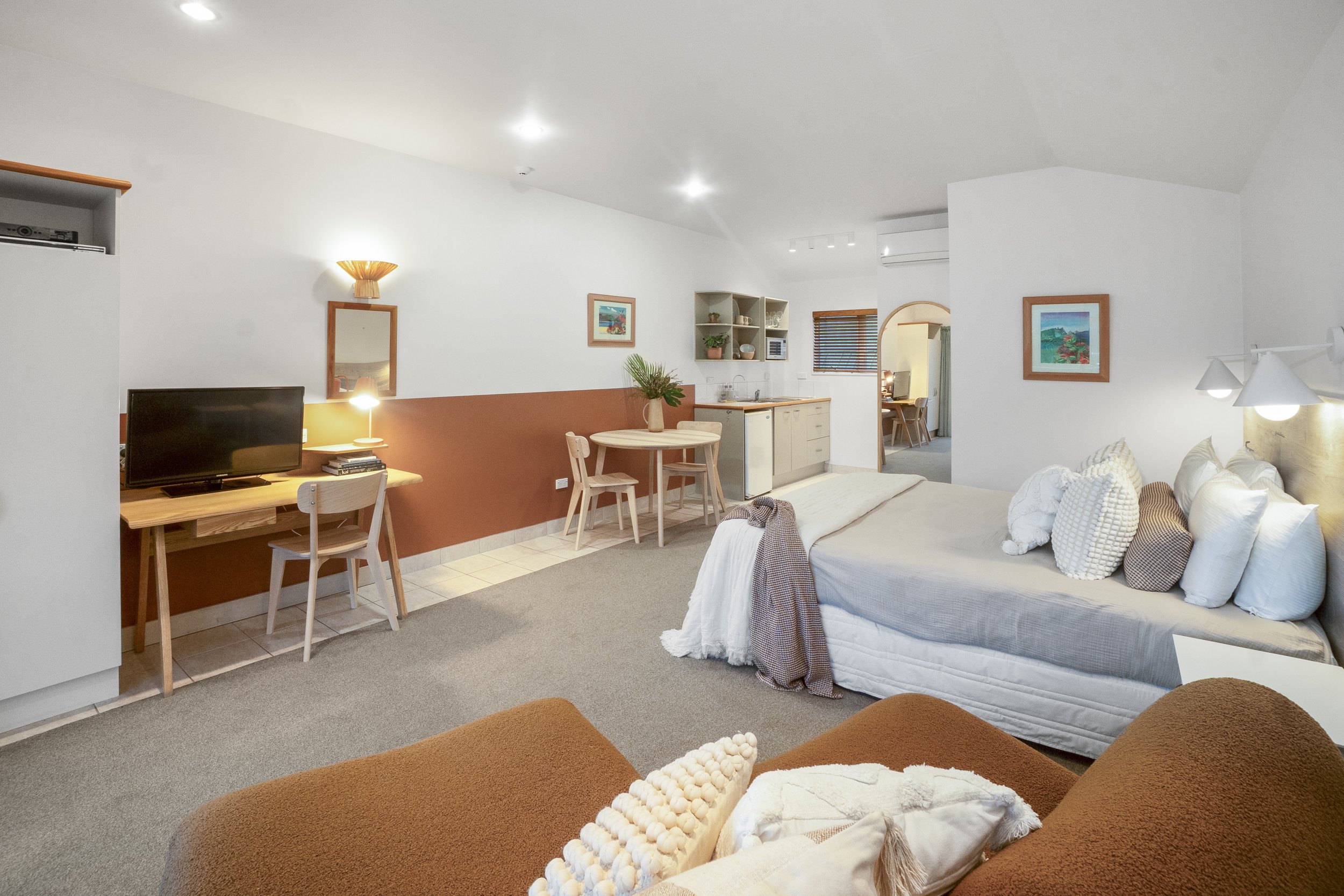 Spacious studio apartment with modern decor, featuring a cozy bed with pillows and blankets, a wooden desk with a TV and chair, a small dining table with chairs, and a kitchenette with cabinets. The room includes a small side table, wall art, and amb