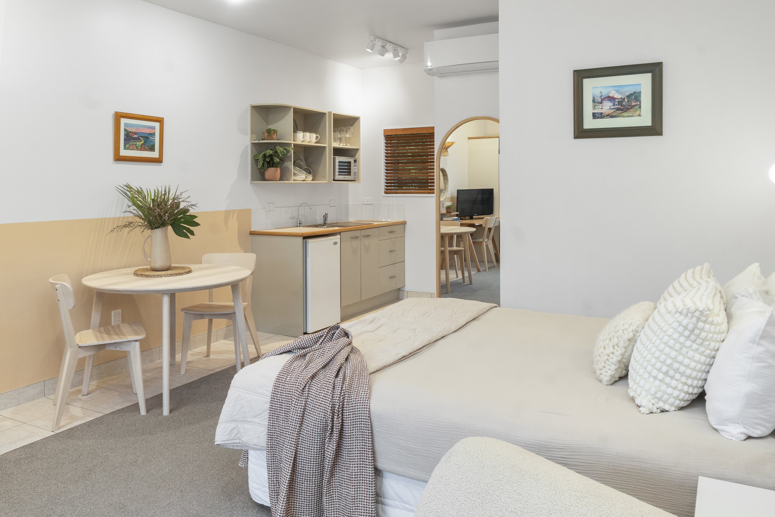 Modern studio apartment interior with a bed, small dining table, kitchenette, and decorative plants.