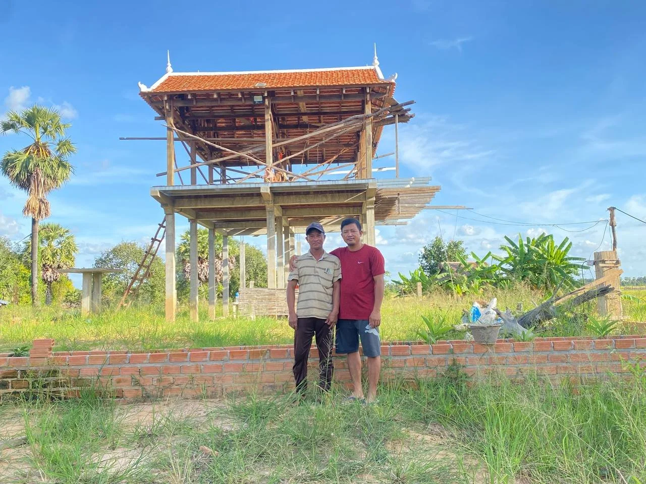 Two men standing in front of a wooden house under construction, with a clear blue sky and green vegetation around.
