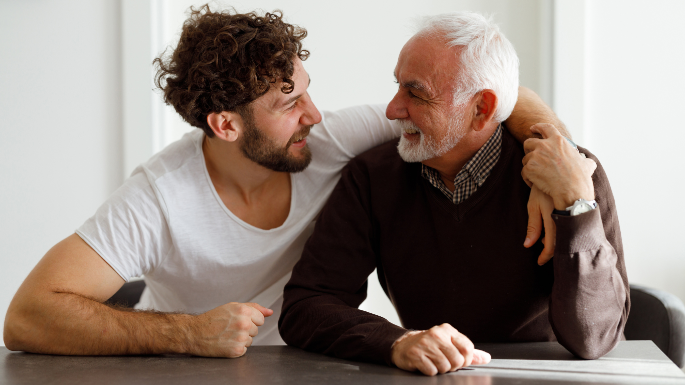 A father and son sat at a table with sons arm around his father and smiling at each other