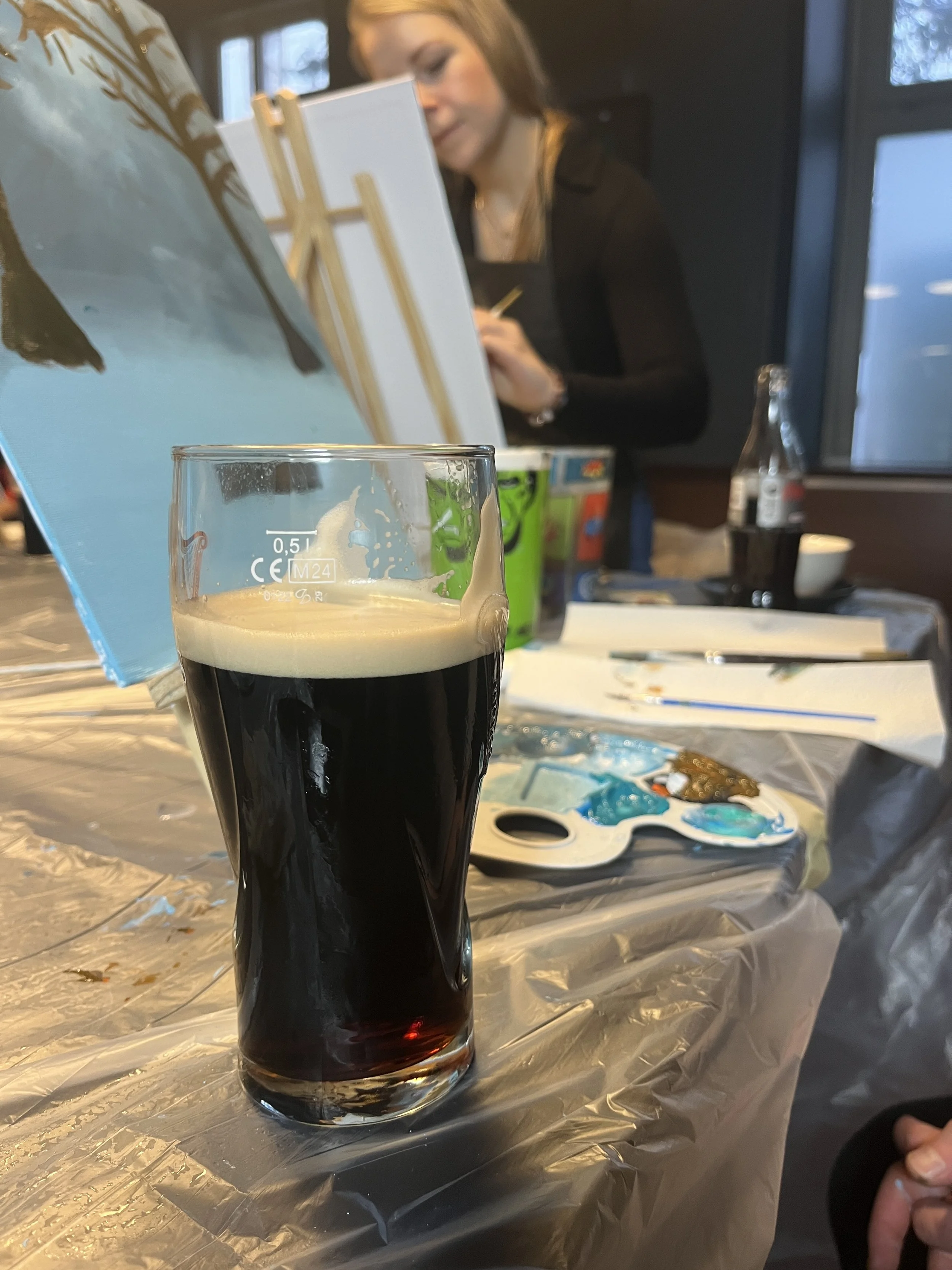 A pint of dark beer with a creamy head on a table covered with a plastic sheet, with an artist painting on a canvas in the background.