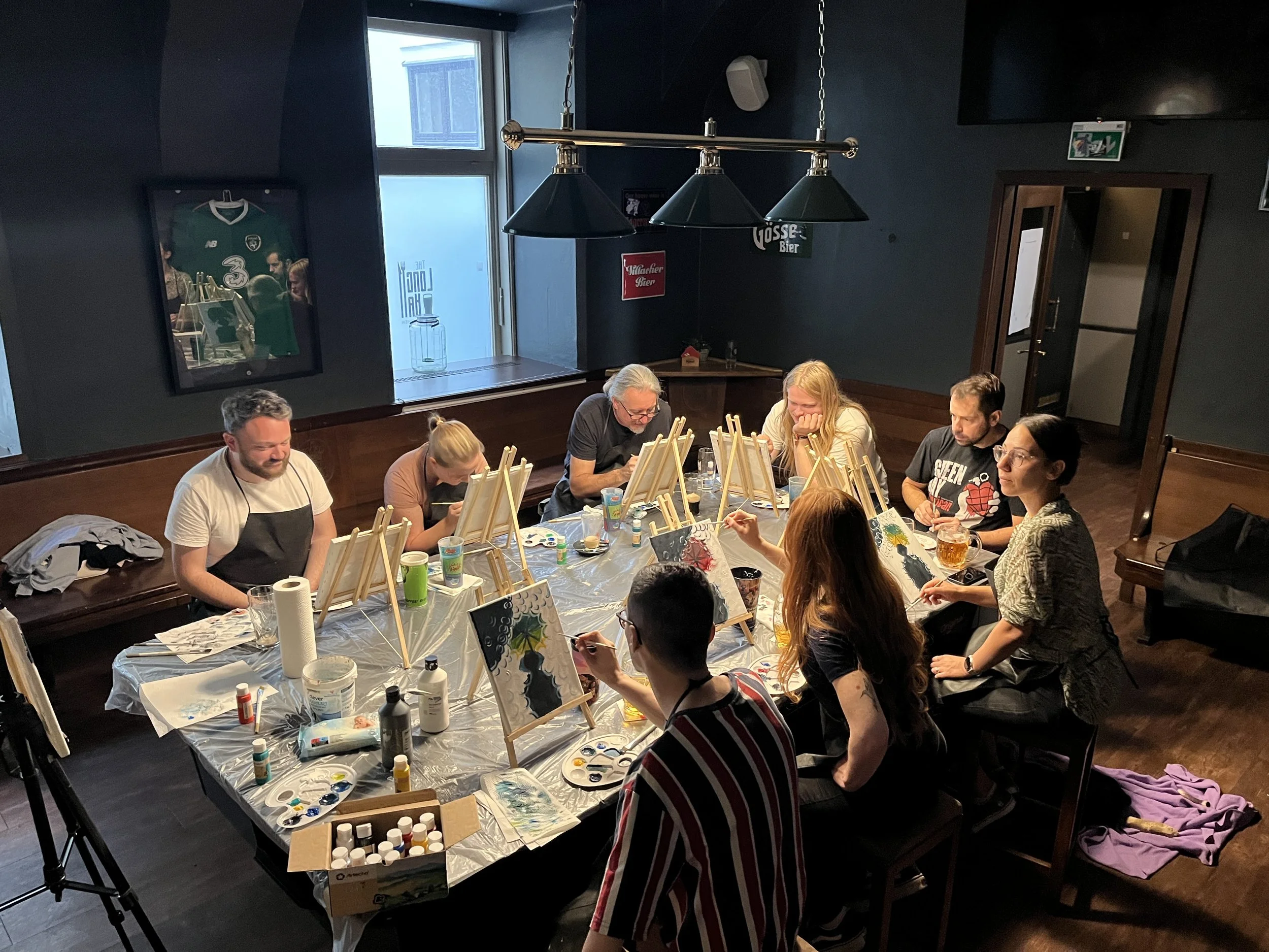 Group of nine people painting on canvases at a table in a dimly lit room, with painting supplies and drinks on the table.