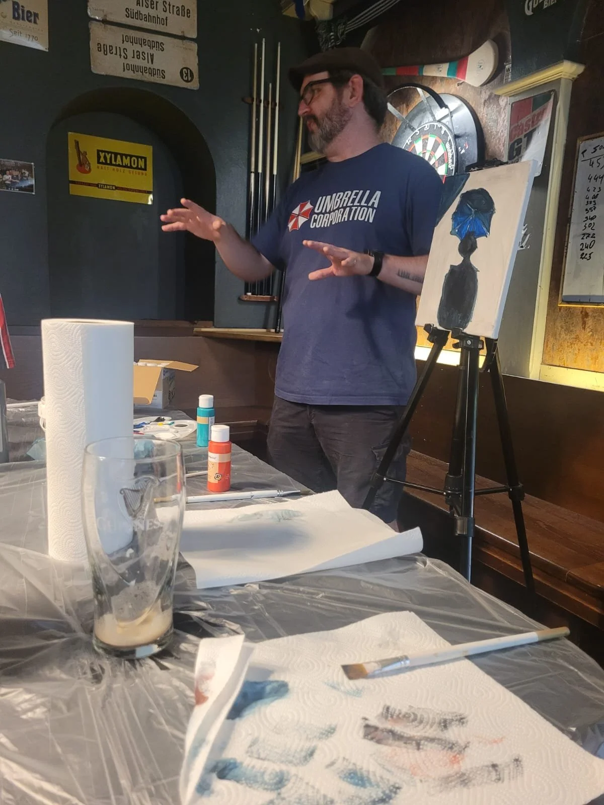 A man wearing glasses and a blue t-shirt with an umbrella logo is giving a presentation or saying something while standing near a painting on an easel. The painting depicts a person with a blue head covering inside a dark bottle. The table in front h
