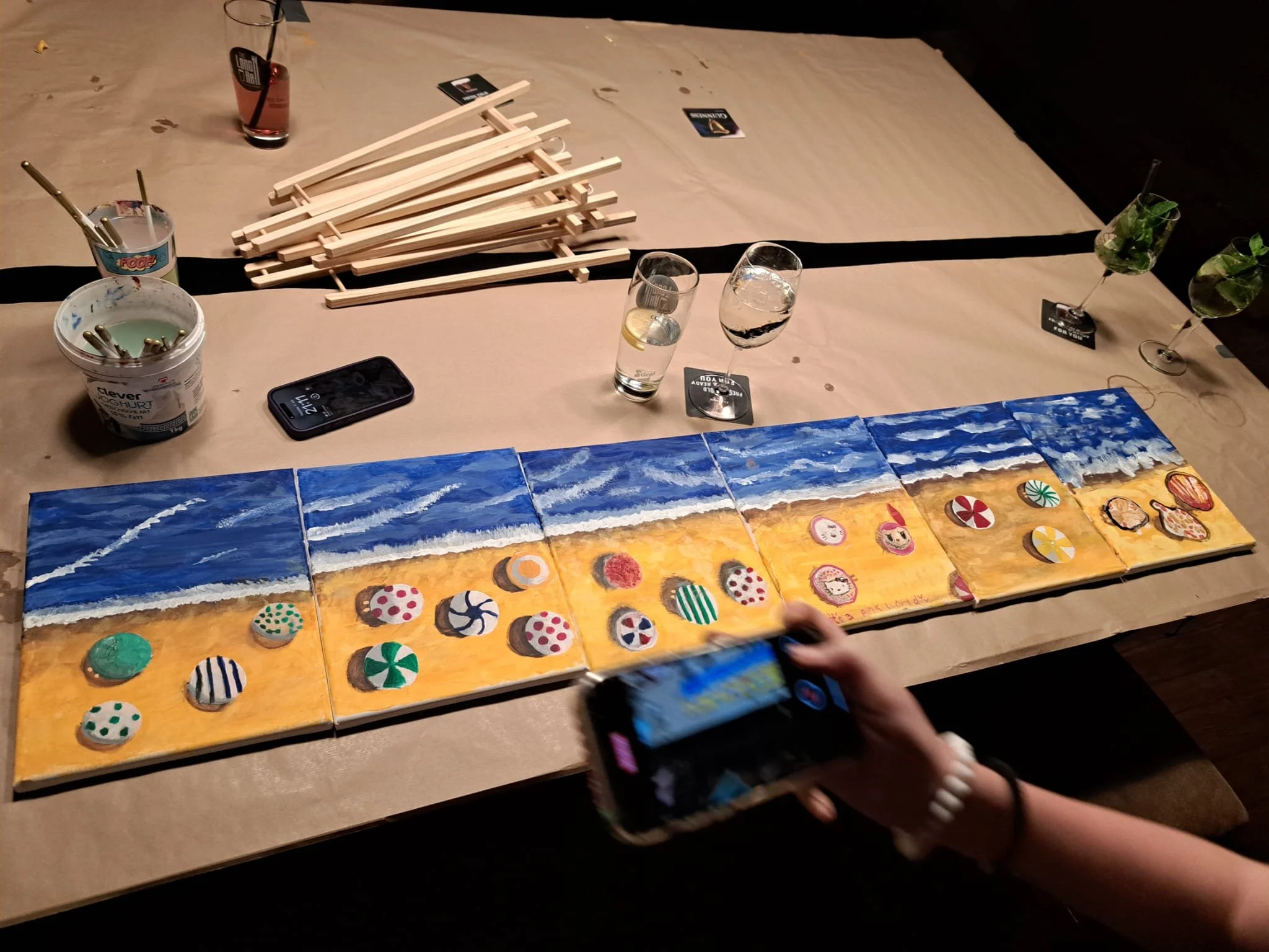 A long table with a beach-themed painting divided into six sections as a centerpiece. The painting shows ocean waves meeting a sandy shore, with decorated cookies resembling beach balls and umbrellas placed on the sand. There are several drinks on th
