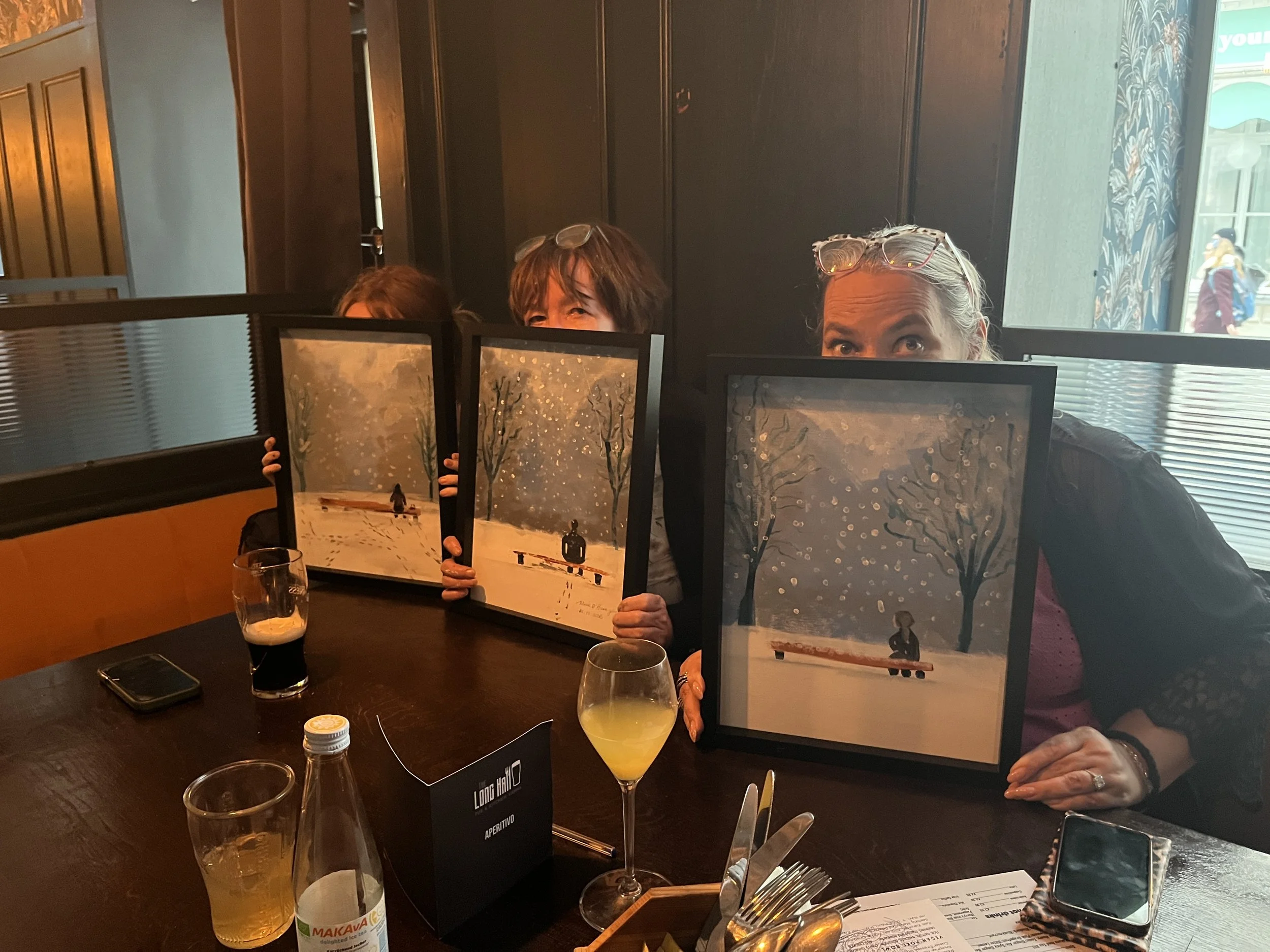 Three women sitting at a table in a restaurant, holding framed artwork with a snowy winter scene. The woman on the right is wearing glasses on her head and smiling, while the woman in the middle and another woman on the left are partially hidden behi