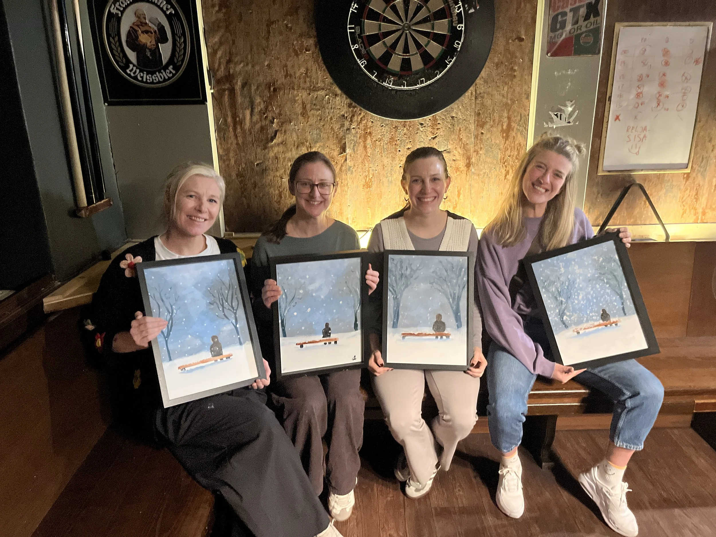 Four women sitting on a wooden bench in a bar or pub, holding framed paintings of a winter scene with a person sitting on a bench in a snow-covered park. There is a dartboard on the wall behind them, along with a whiteboard and some signs.