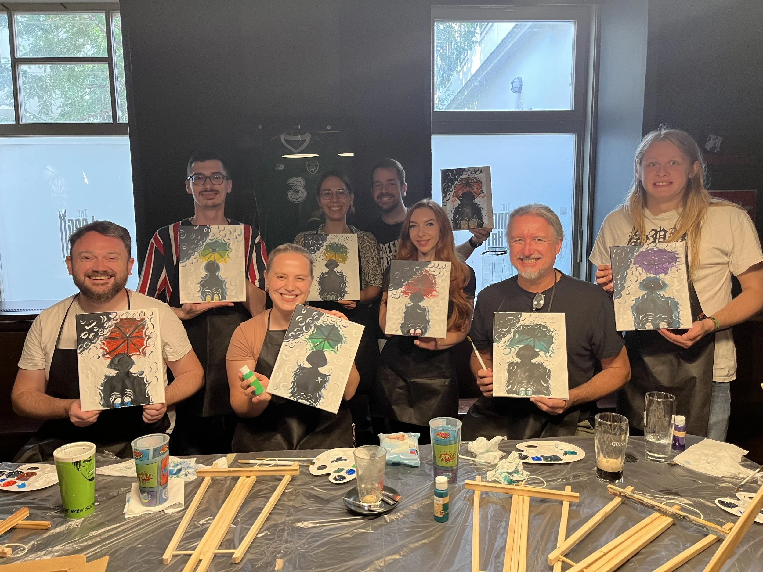 Group of people participating in a canvas painting activity indoors, holding up their completed paintings of a silhouette holding an umbrella with stormy clouds background.