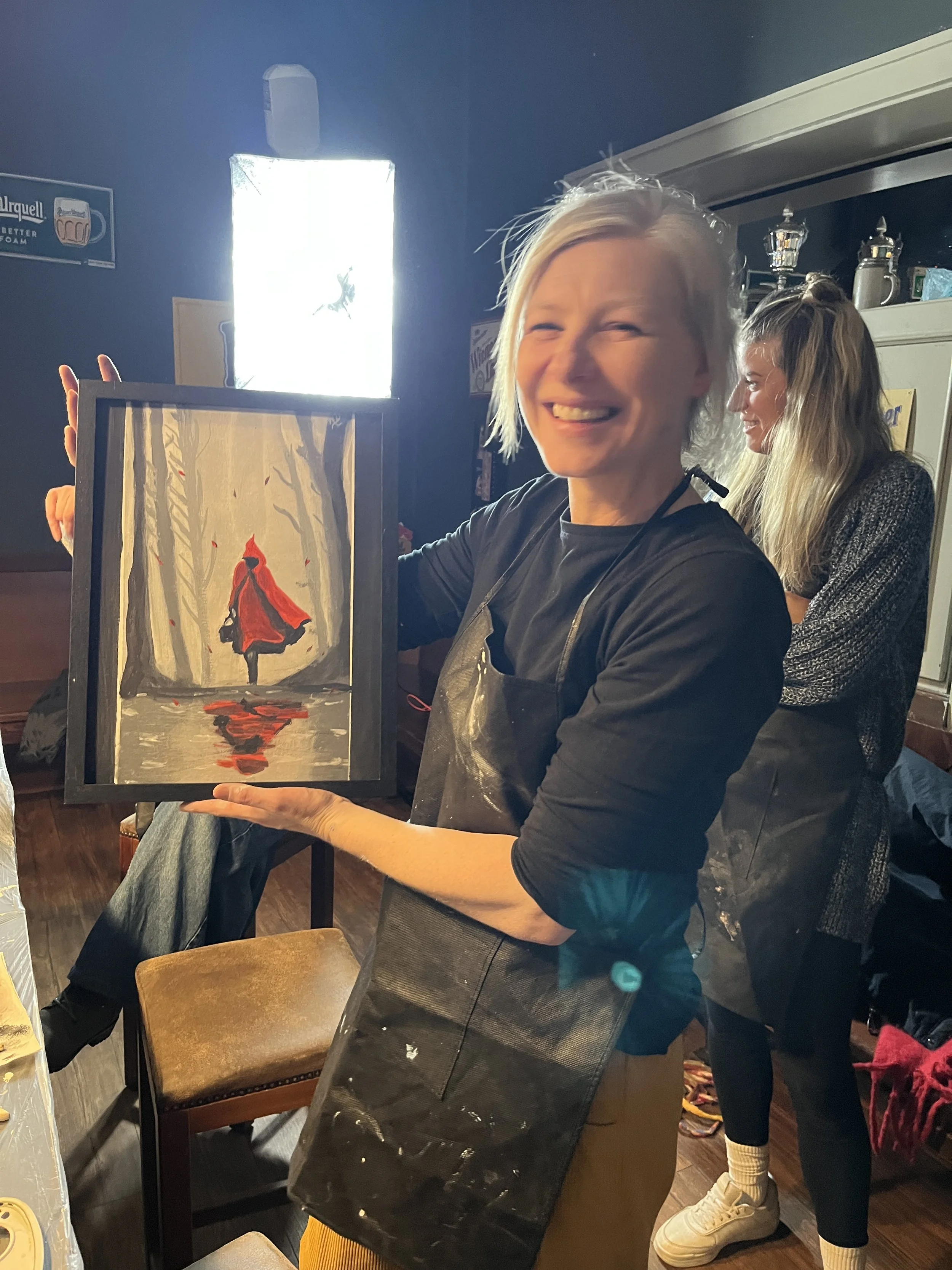 Woman smiling and showing a painting of a figure in a red cloak walking in a forest, with a reflection of the figure in water, during an art activity in a cozy room.