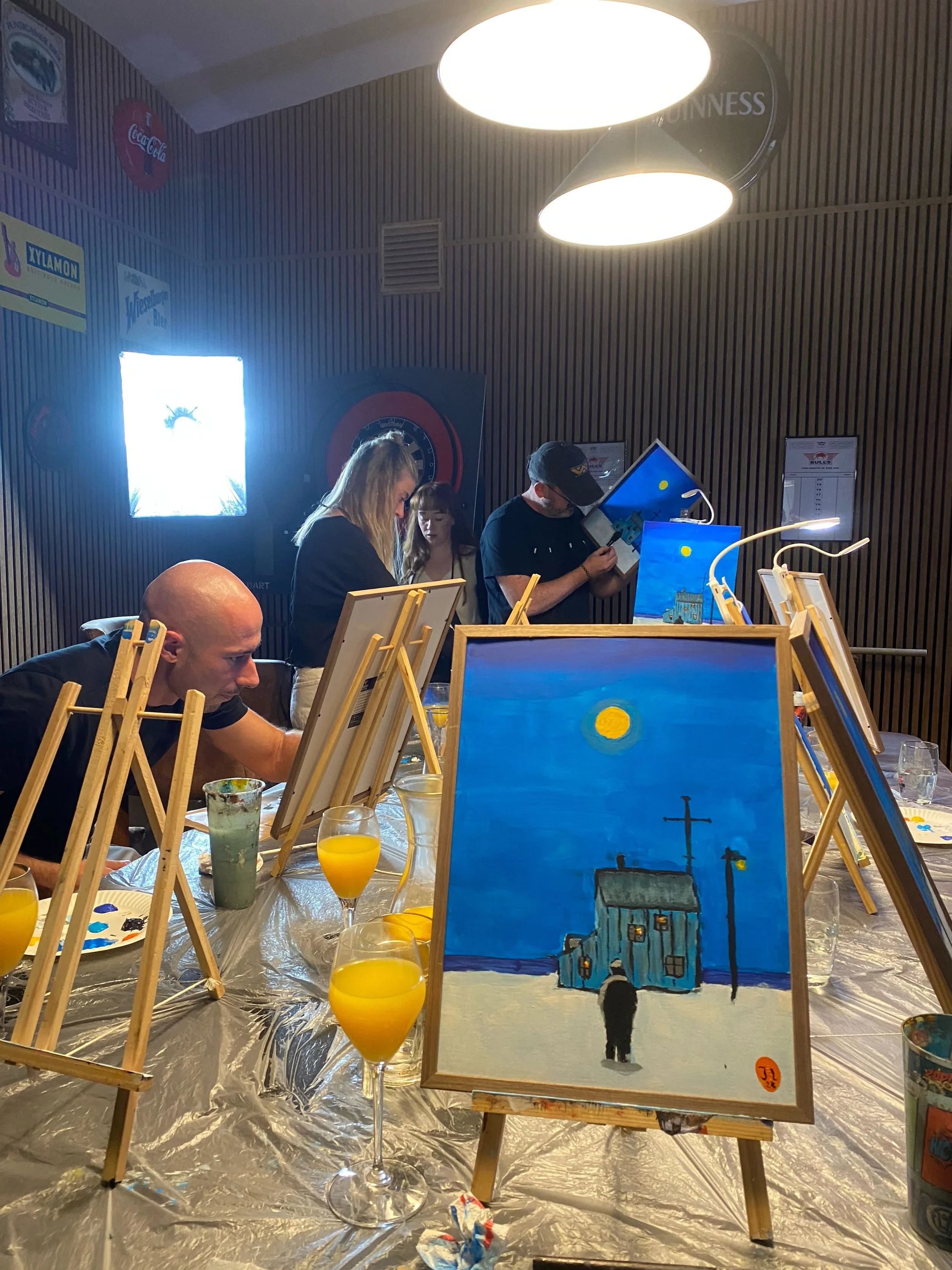 People painting and viewing artwork in an art class with paintings of buildings and a night scene on display.