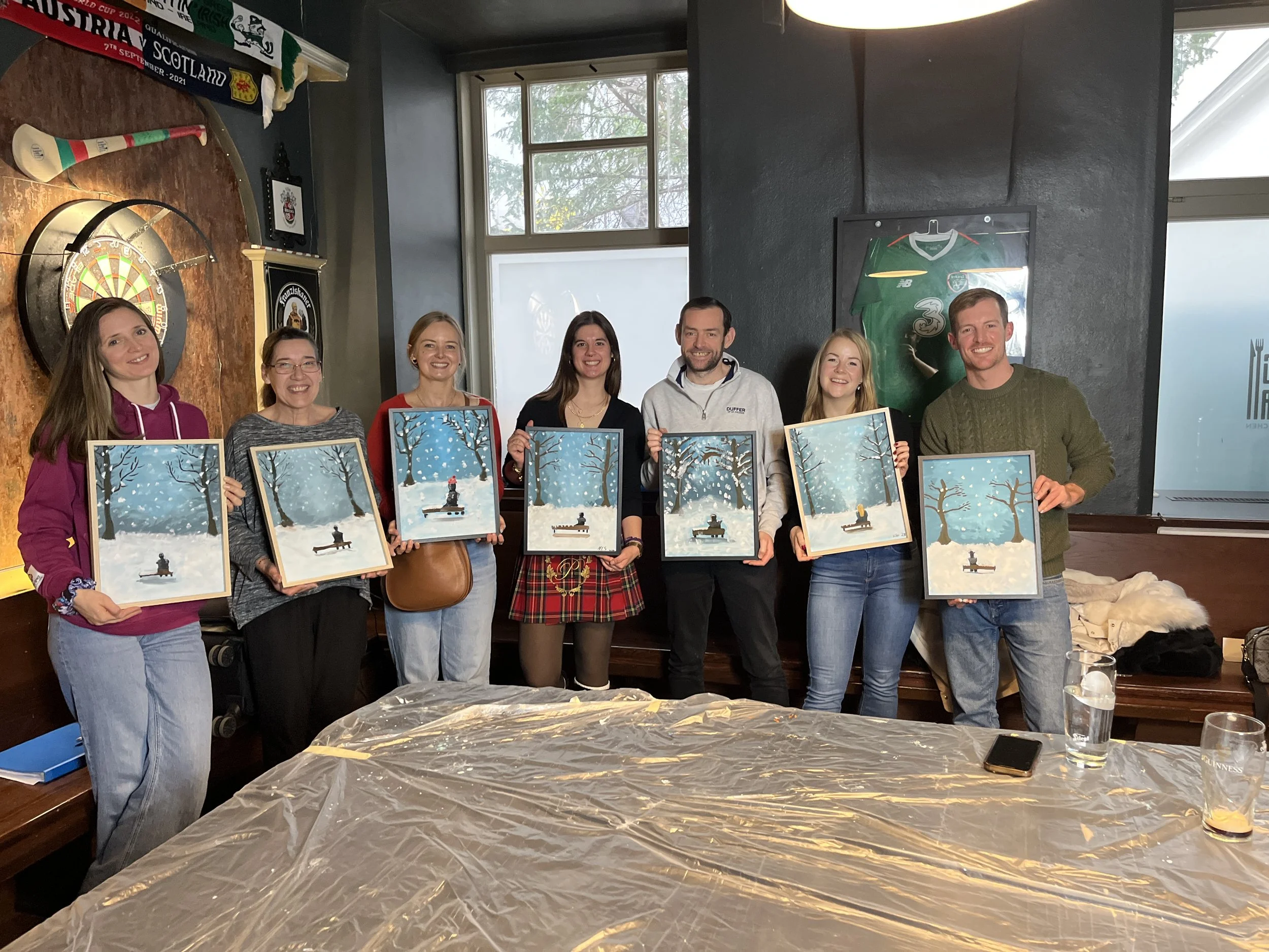 Group of seven people holding framed snow-themed paintings inside a room with dark walls, a dartboard, and rugby jerseys on display, with a table covered in clear plastic in the foreground.