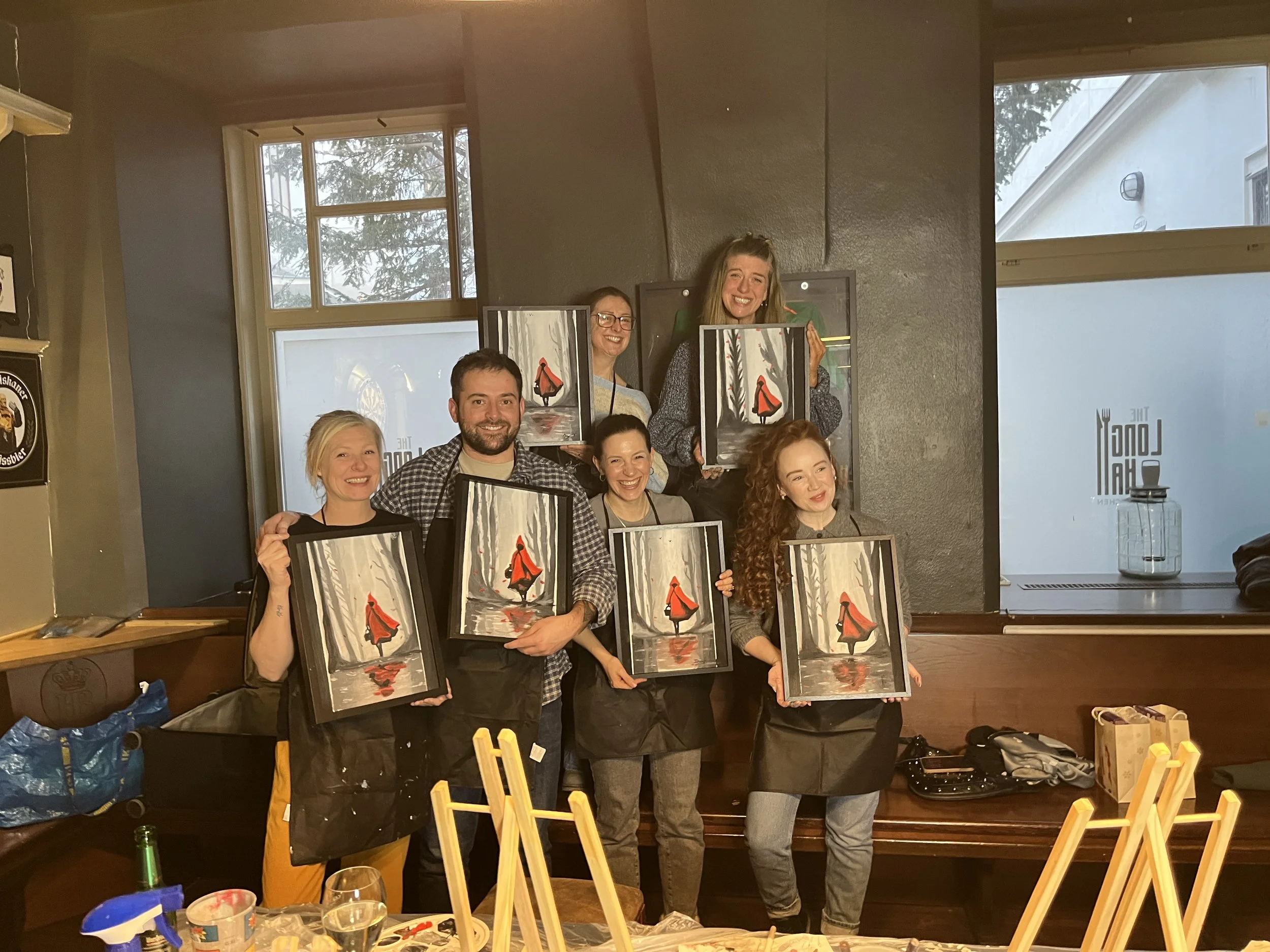 Group of six people smiling and holding paintings of a girl in a red hooded cloak walking through a forest with tall trees, inside a room with large windows.