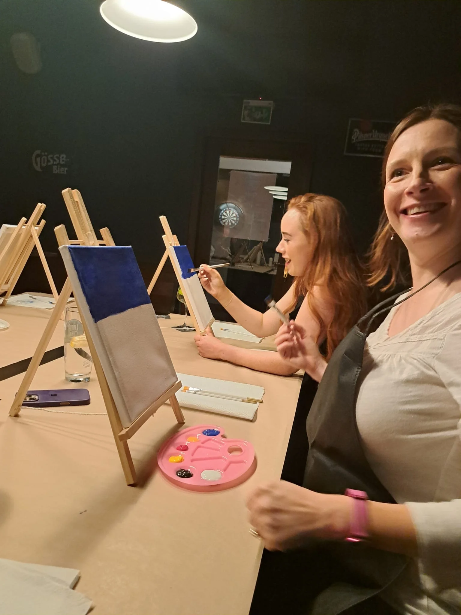 Women painting on small canvases at a paint and sip event in a dimly lit room.