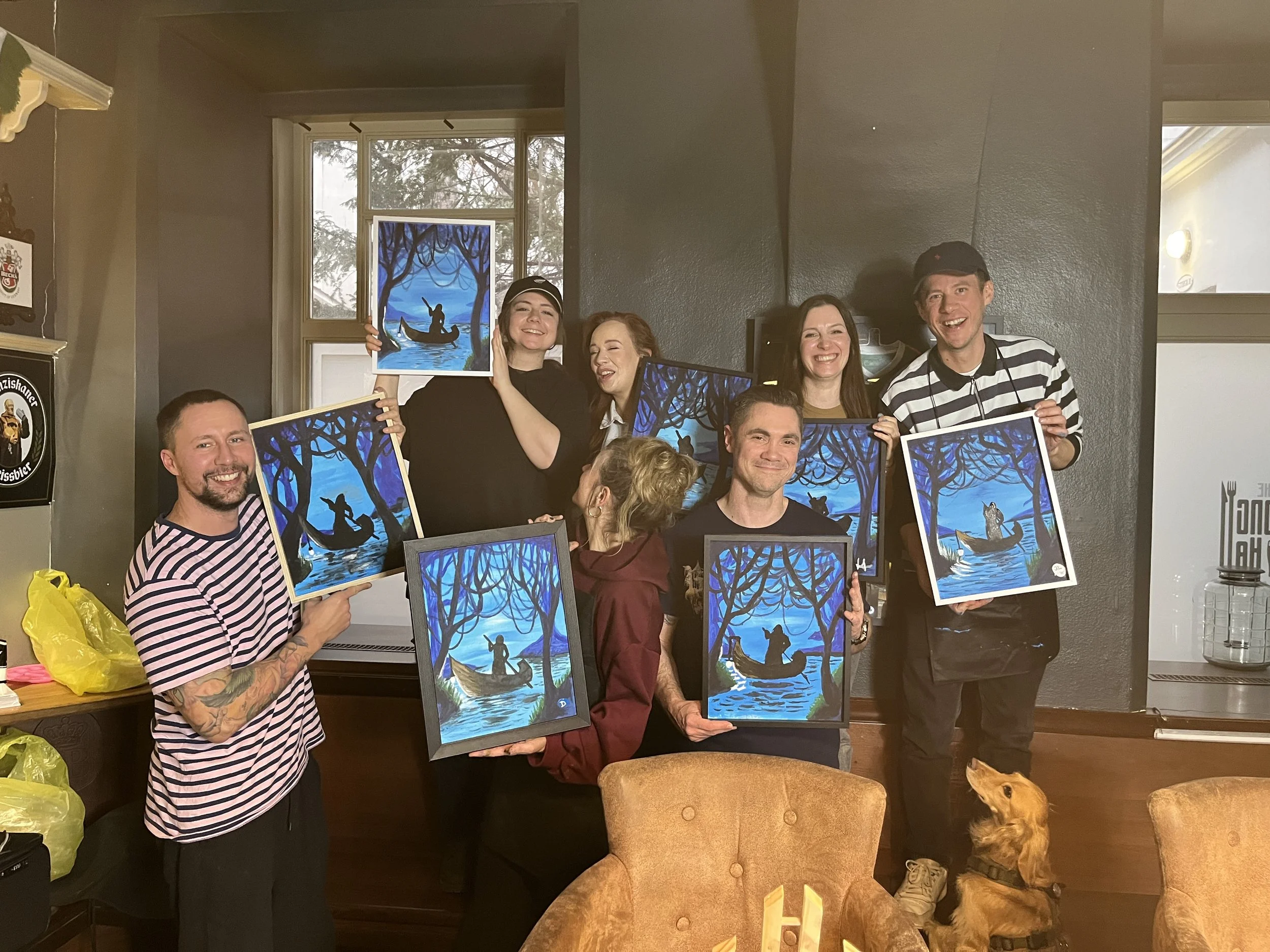 Group of people holding paintings of a person in a boat on a lake at night, surrounded by trees, in a cozy indoor setting. A dog is sitting on the floor in front of one of the chairs.