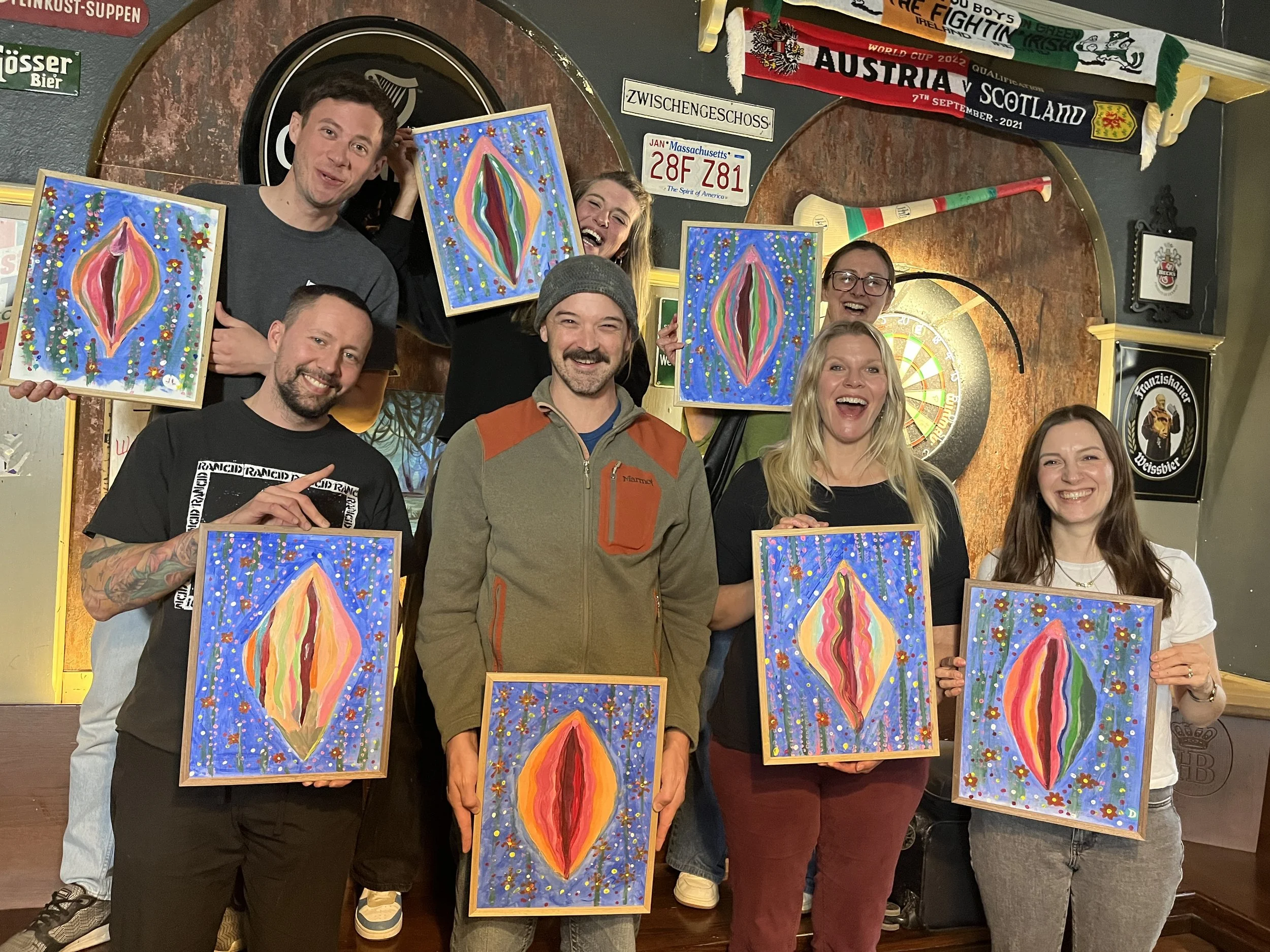 A group of eight friends in a bar, smiling and holding colorful abstract paintings. The background features a dartboard, flags, and signs, with a lively and cheerful atmosphere.