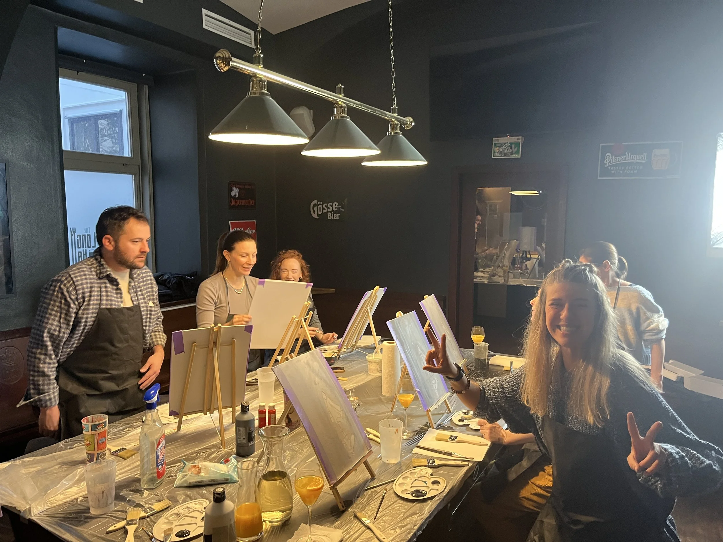 Group of people participating in a painting class in a dimly lit room, with blank canvases on easels, art supplies on the table, and a cheerful woman smiling and posing with peace signs.