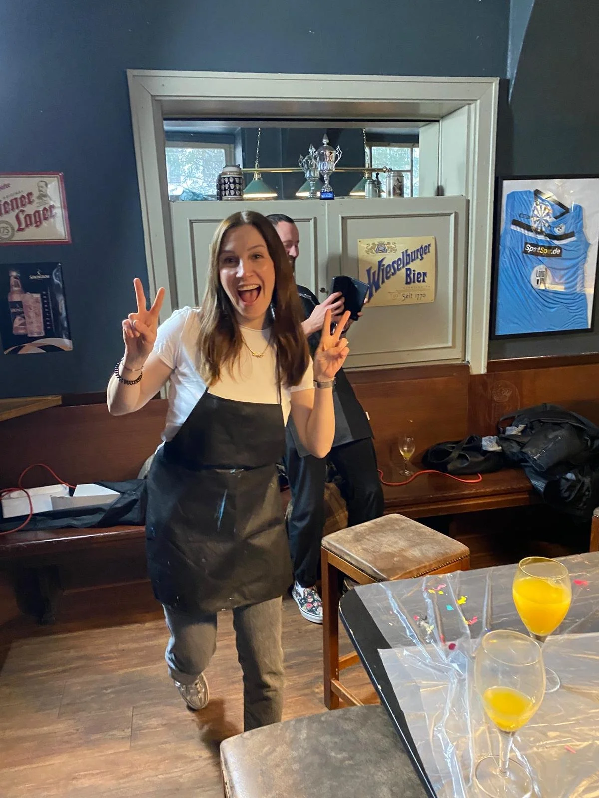 A woman smiling and making peace signs with both hands in a restaurant or bar setting, with another person behind her holding a phone and two glasses of orange juice on a table in the foreground.
