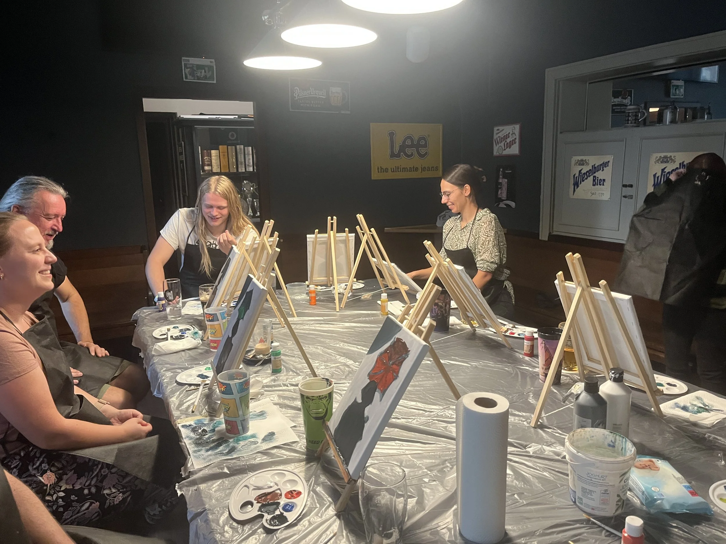 People participating in a group painting activity, sitting around a table covered with a plastic sheet, with several small canvases on easels and painting supplies. They are smiling and engaging in painting in a dimly lit room decorated with posters.