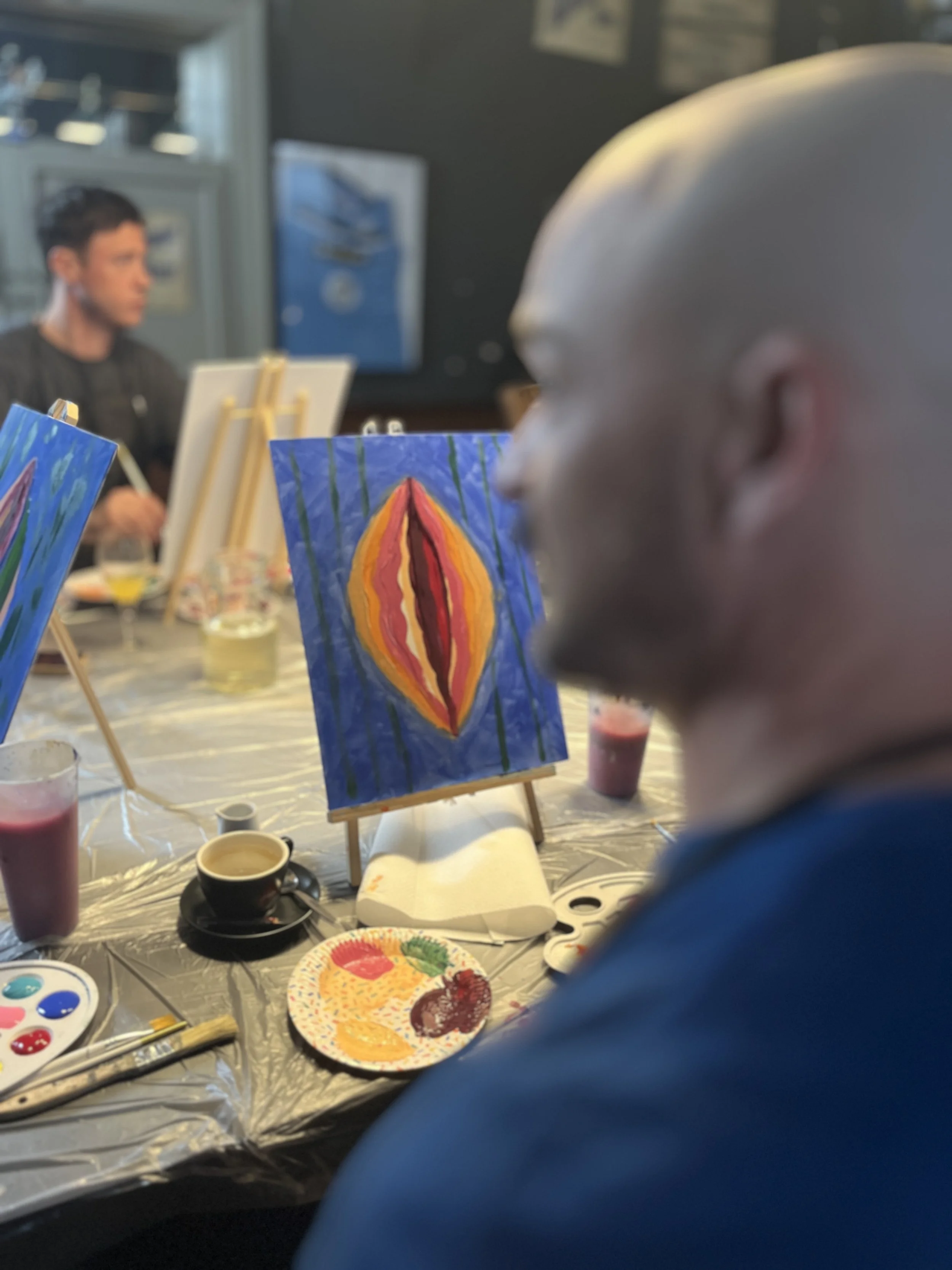 Person with a bald head in profile view painting a colorful abstract artwork featuring a bright red, orange, and pink shape on a blue background, sitting at a table with painting supplies during an art class.