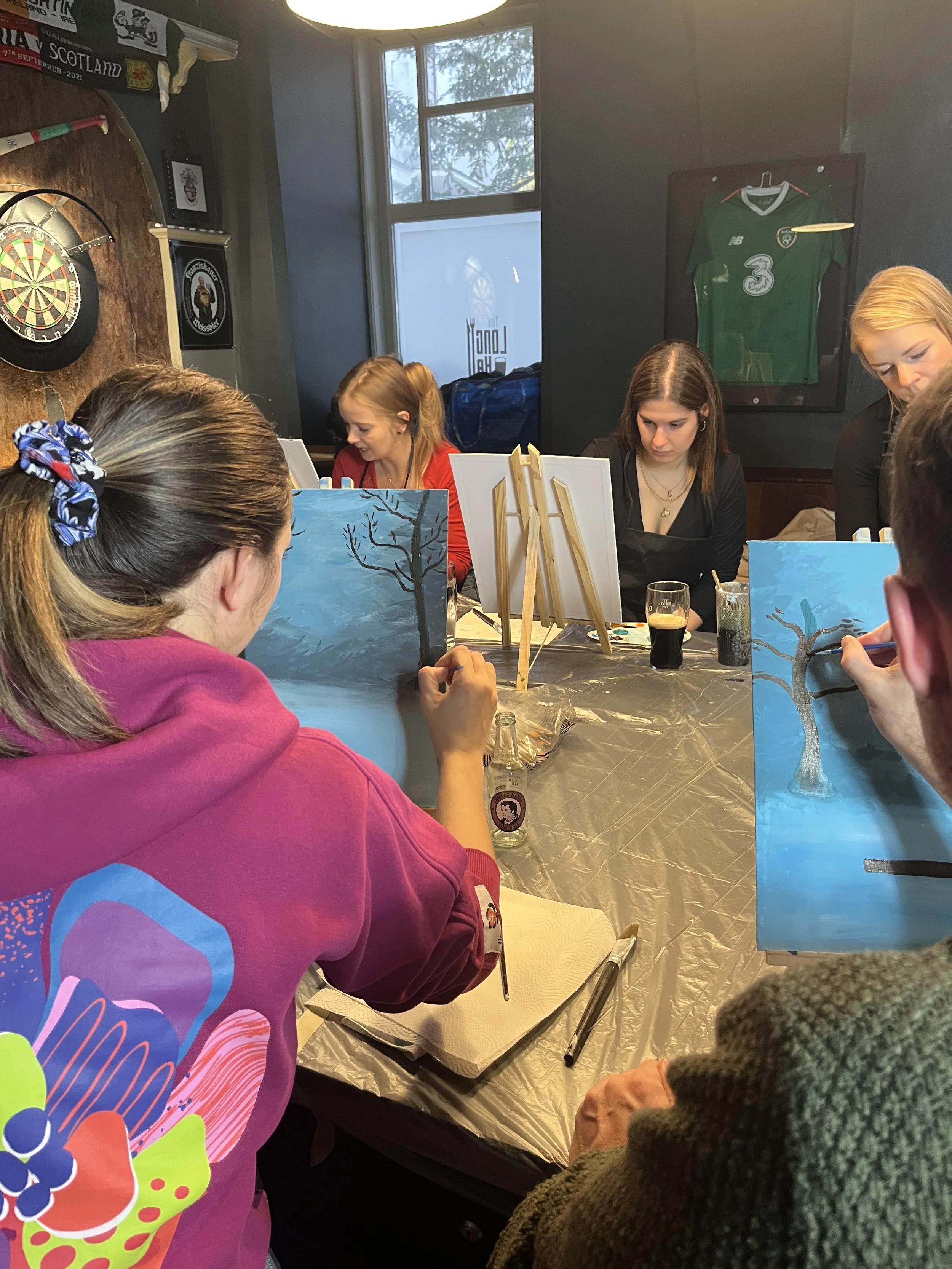 A group of people painting landscape pictures of trees on canvases at a table in an art studio. Two women are focused on their canvases, with one in a pink hoodie and another in a dark sweater. There are drinks and painting supplies on the table, and