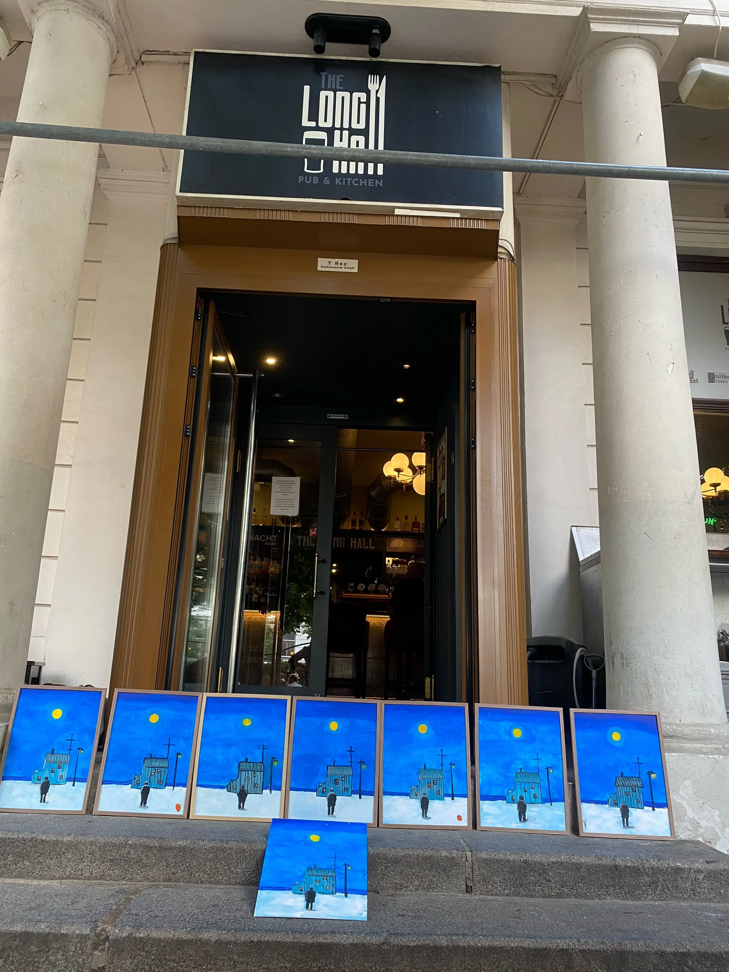 Paintings of a person walking past a blue house under a yellow sun on a blue sky, displayed outside a building with a sign reading "The Long Shot Pub & Kitchen."