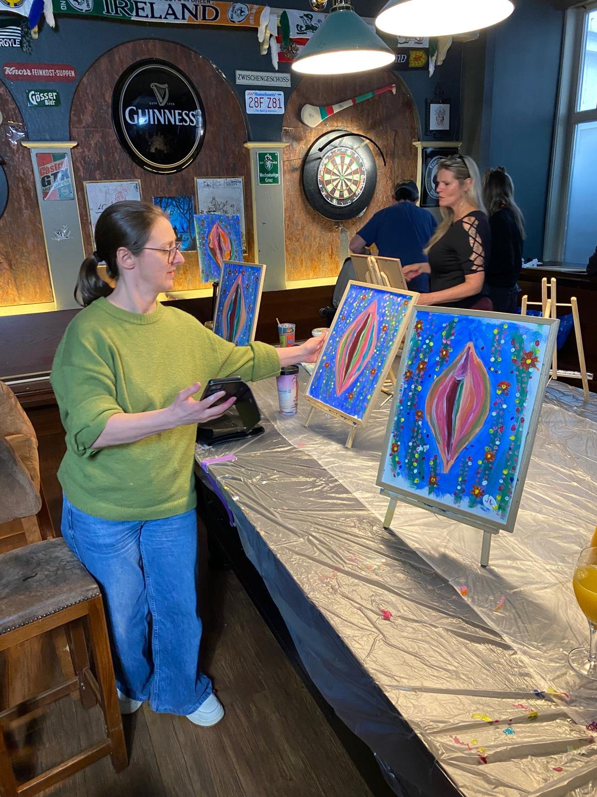 A woman in a green sweater and blue jeans looks at her phone while standing near a table with three colorful abstract paintings on small easels. The table is covered with a plastic sheet, and there are paint supplies and drinks on it. In the backgrou