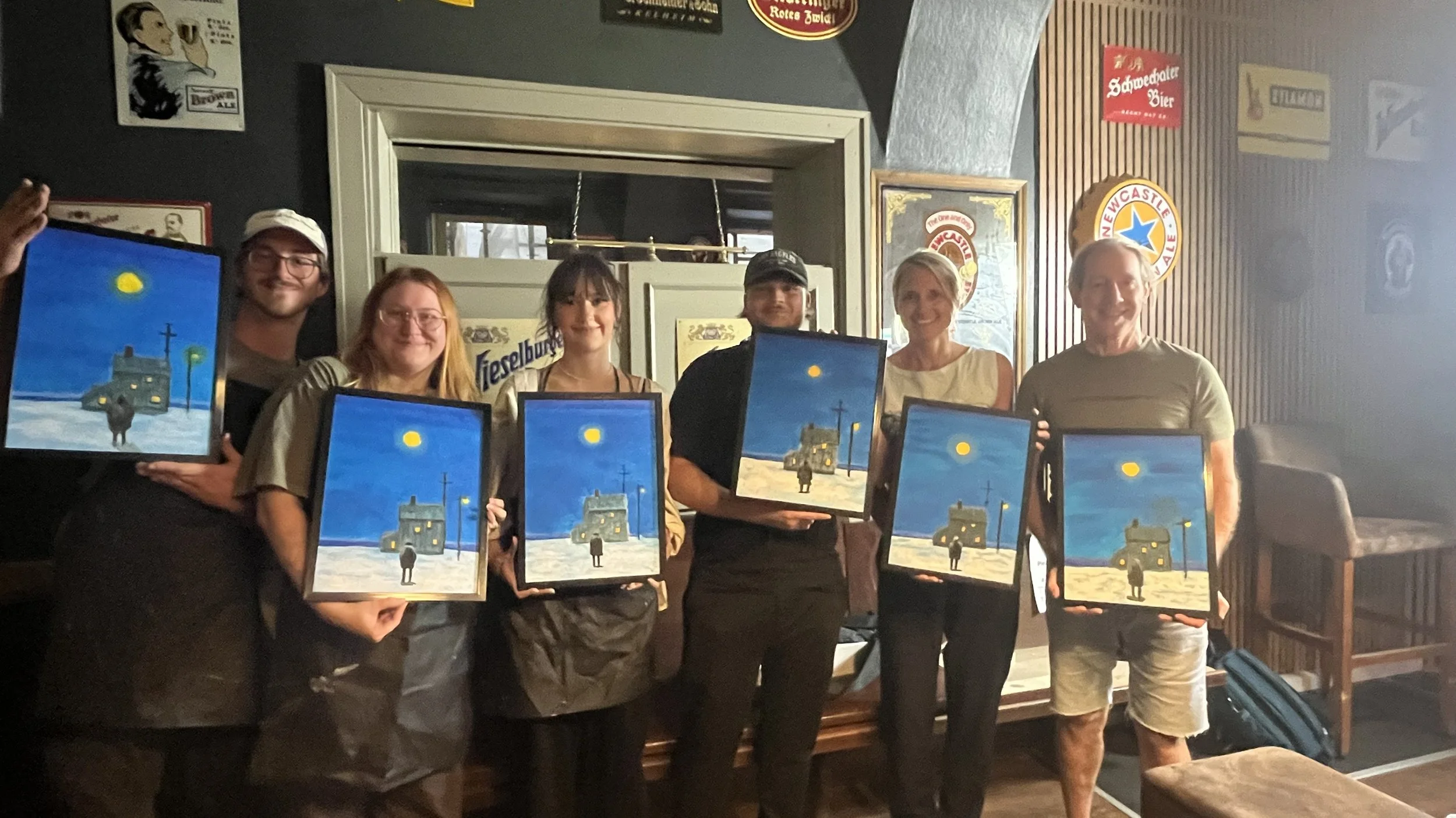 Group of seven people standing in a room, each holding a framed painting of a blue sky, a bright moon, and buildings with lit windows, smiling at the camera.