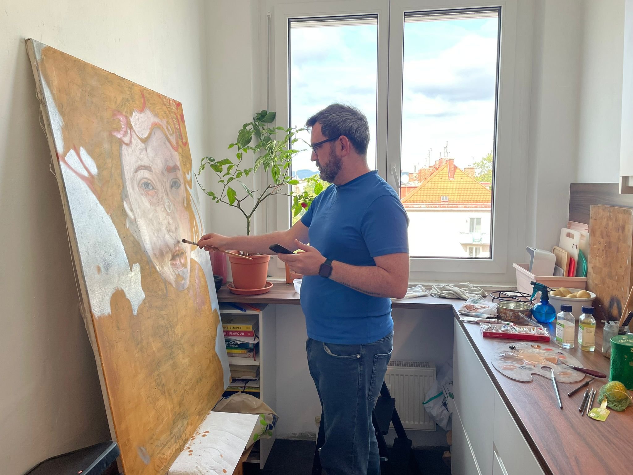 A man painting a large portrait of a woman on a canvas in a bright kitchen with a window in the background.
