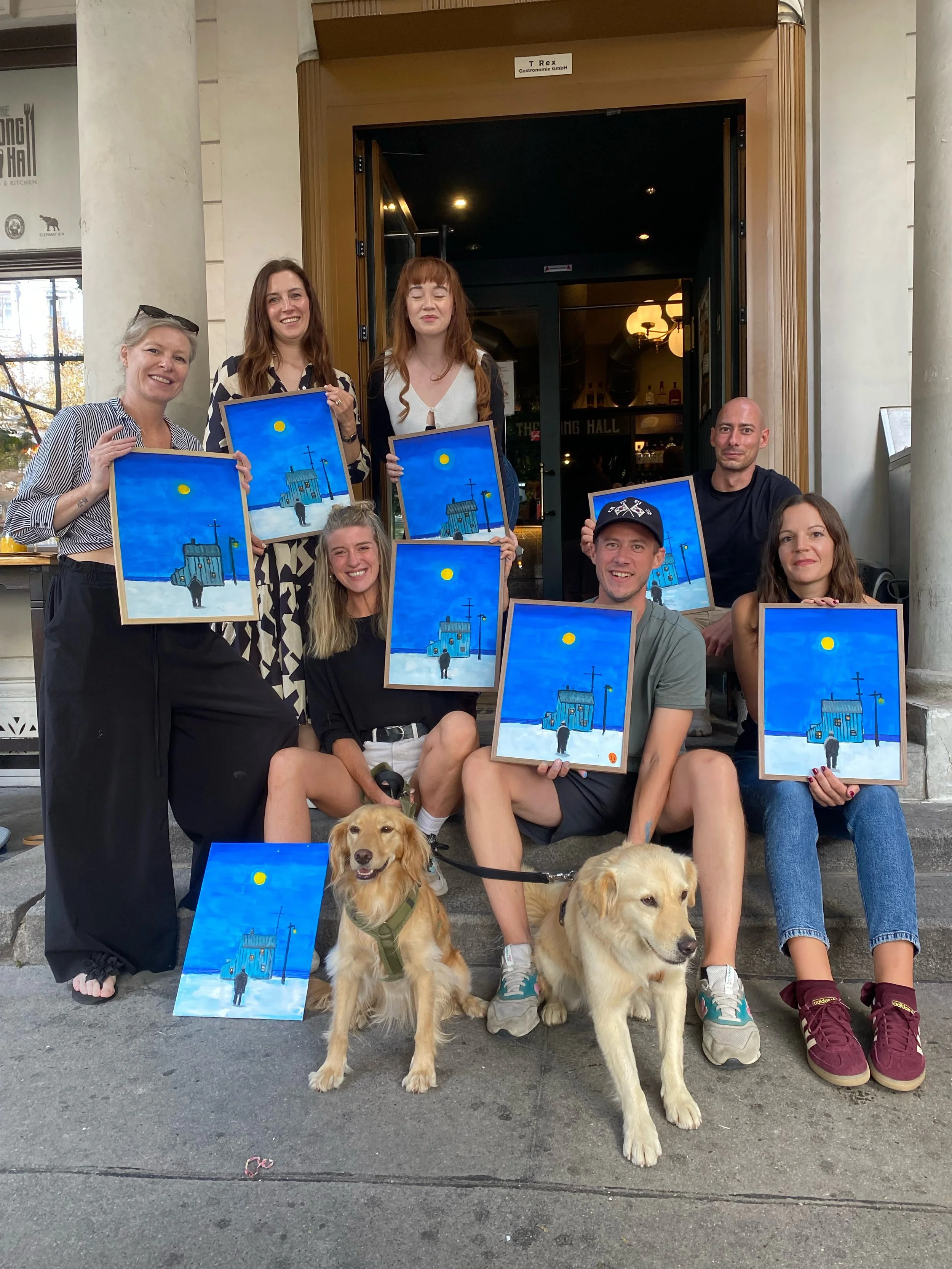 A group of people sitting on the steps outside The Long Hall, Vienna, after a painting and pints session,  holding paintings that depict a blue church and a person walking under a yellow moon. 