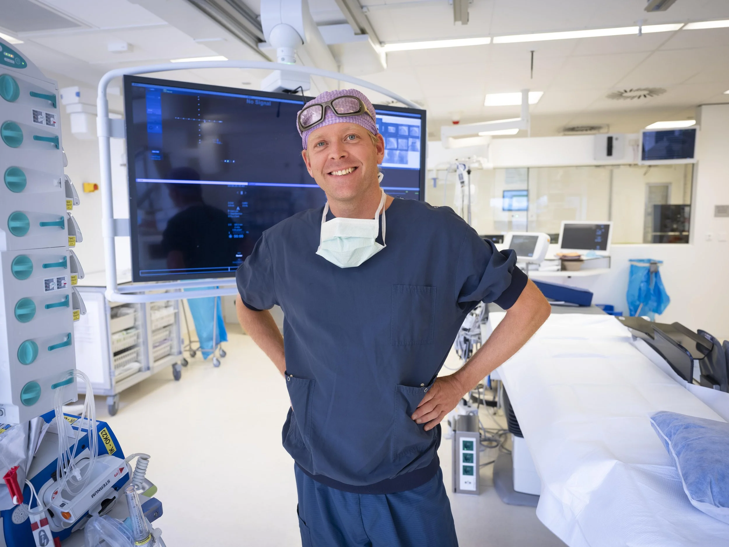 A smiling male doctor or surgeon standing in a hospital operating room, wearing scrubs, a face mask hanging around his neck, and protective goggles on his head, with medical equipment and monitors behind him.