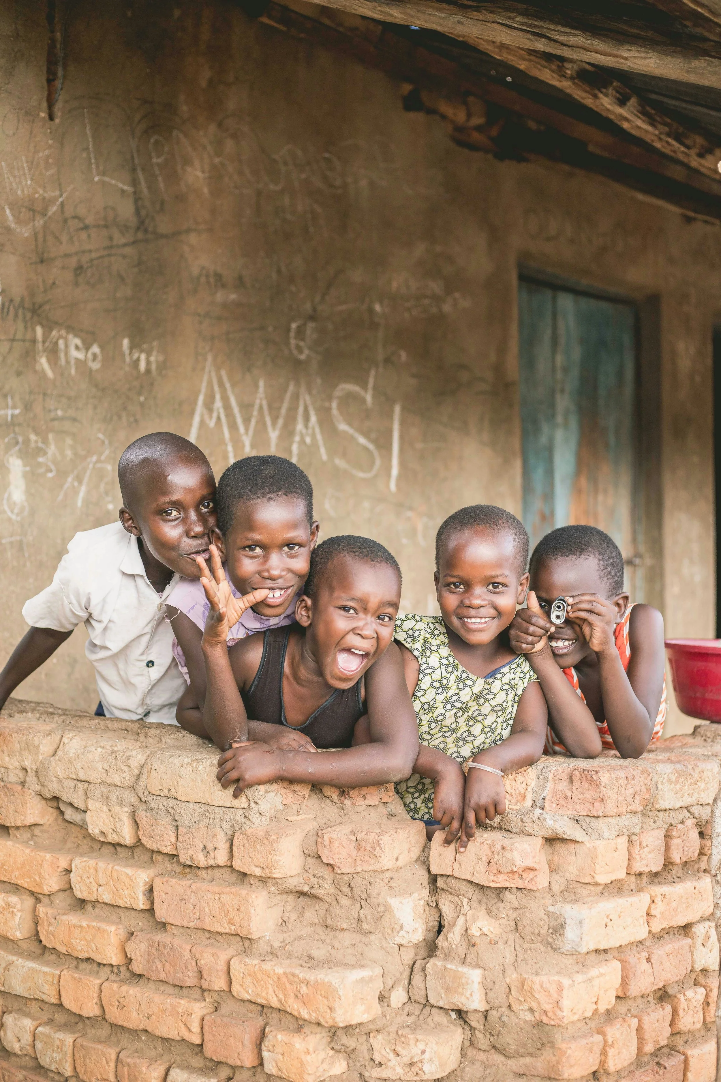 Five children smiling and playing behind a brick wall in front of a weathered wall with chalk drawings and writing, in a rural setting.