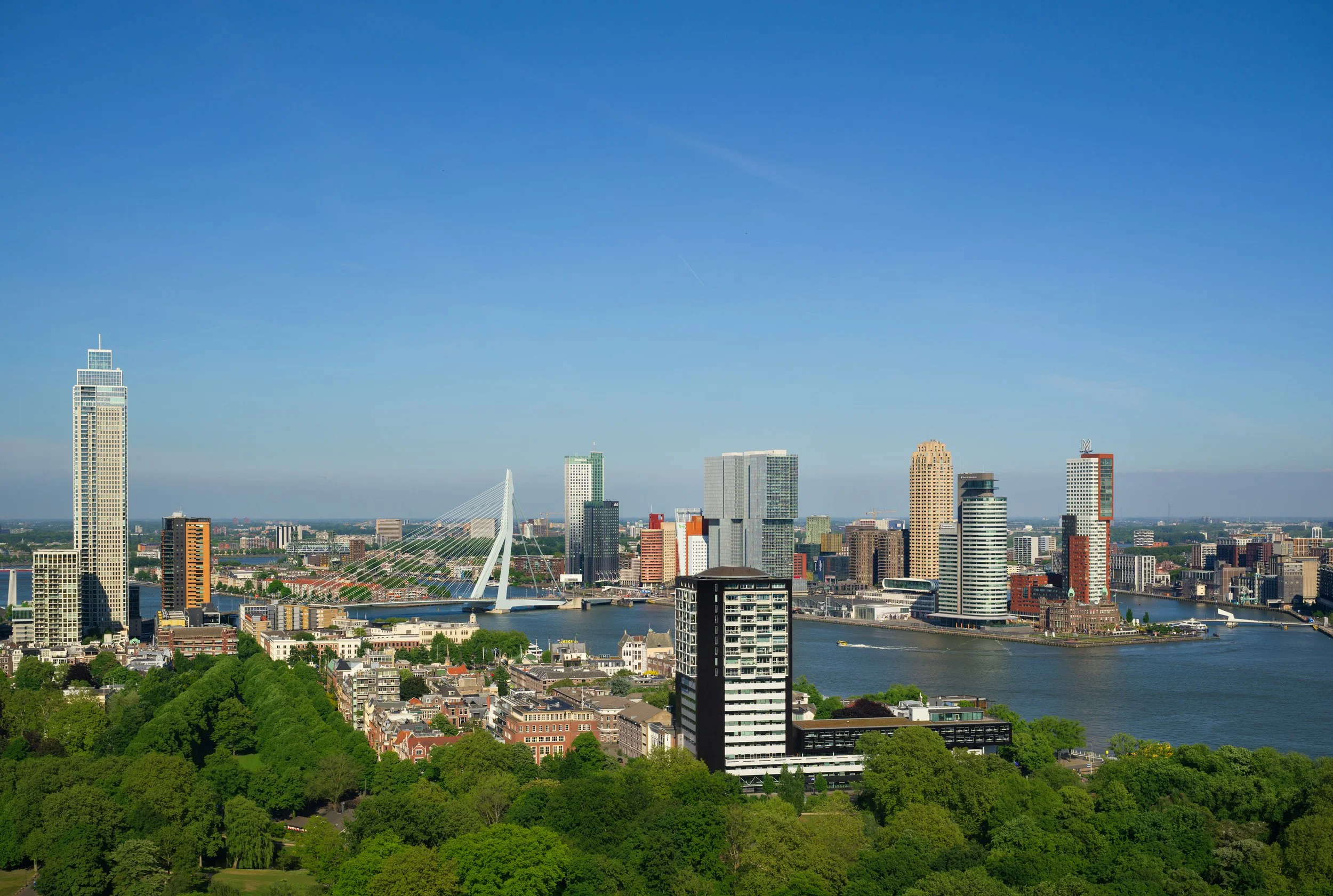 A city skyline featuring tall skyscrapers, a river with boats, and a bridge against a bright blue sky, surrounded by green trees and low-rise buildings.