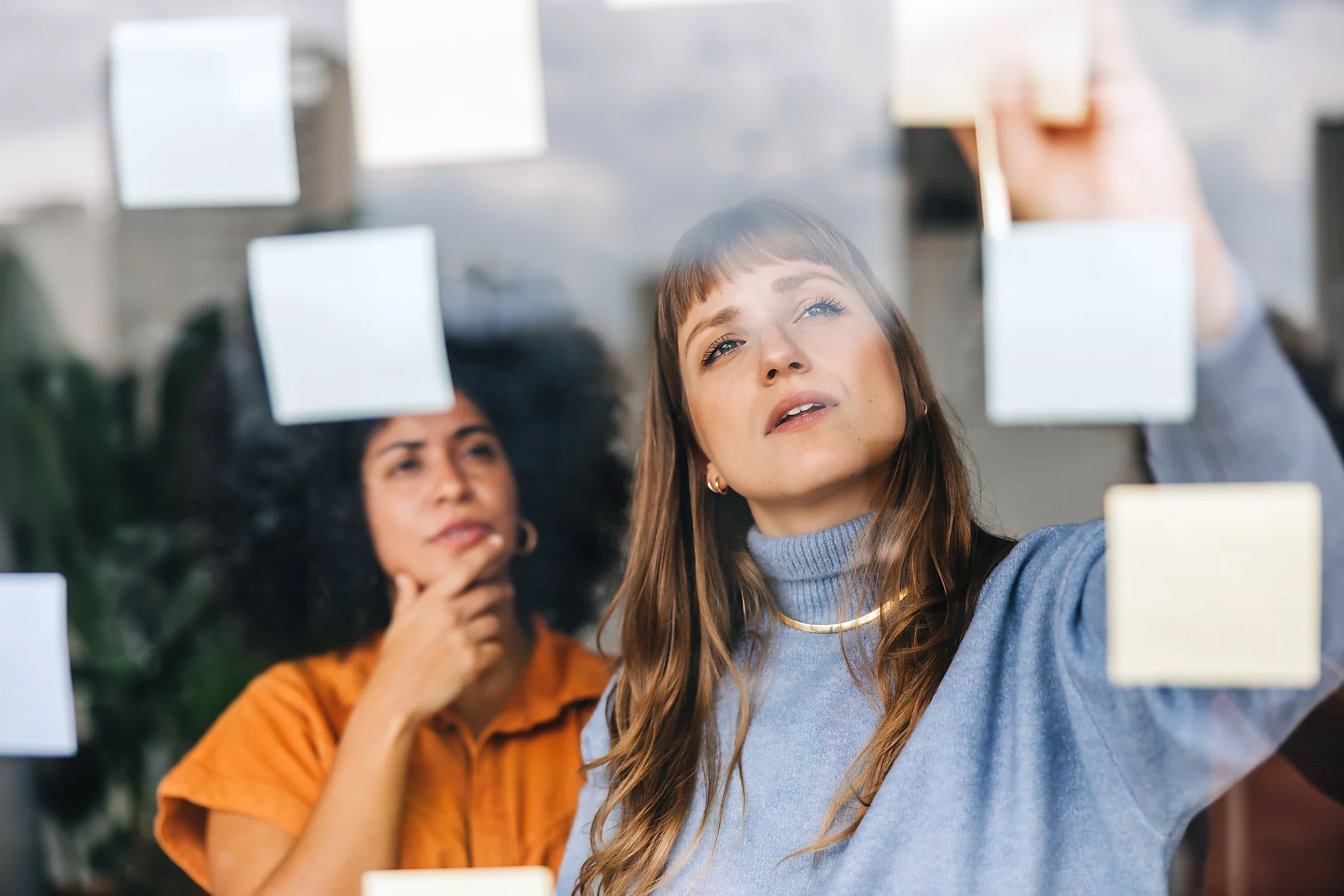 Two women brainstorming in an office, one writing notes on a glass wall with sticky notes while the other observes, deep in thought.