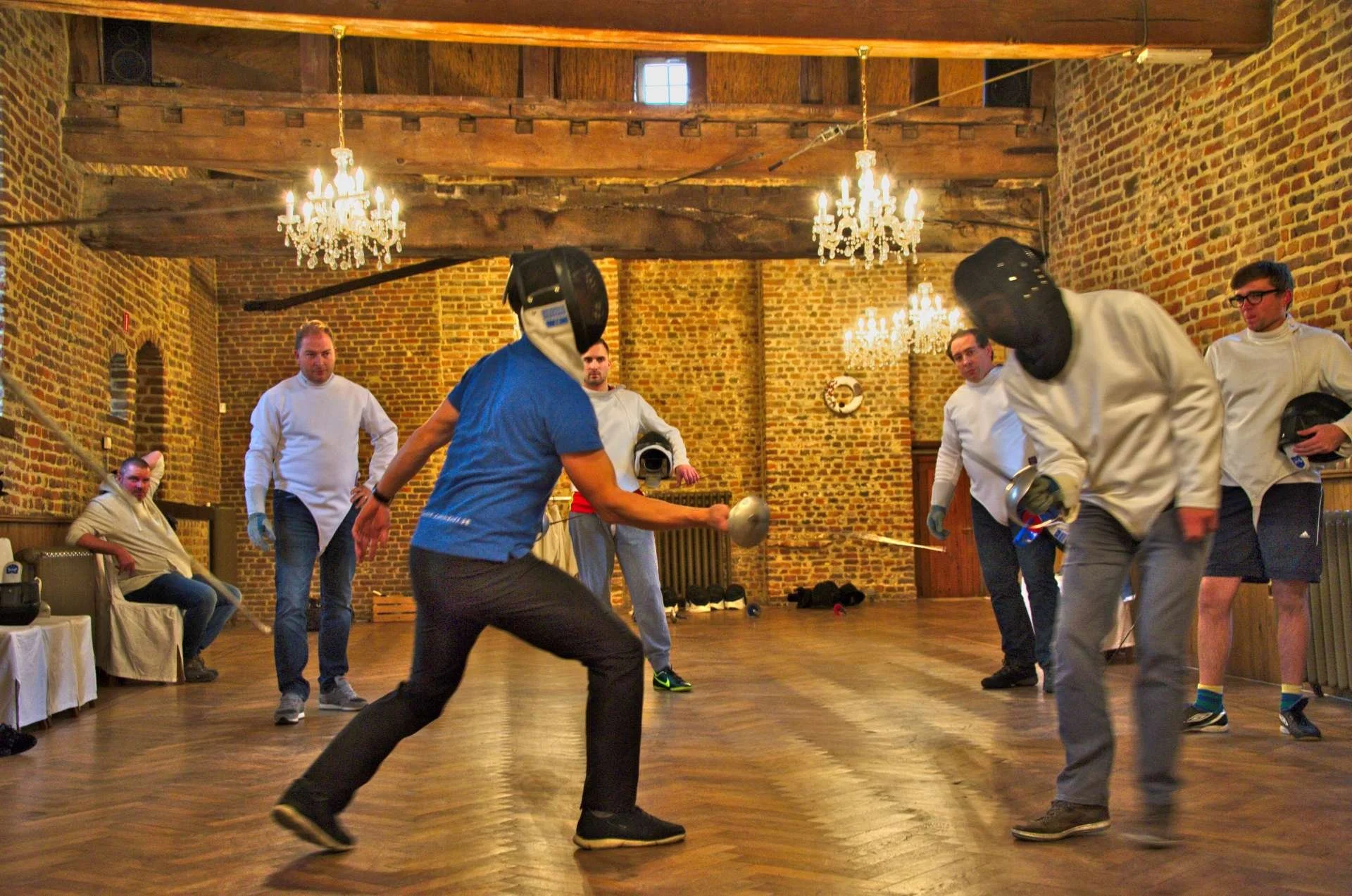 Mensen die schermsport oefenen in een grote zaal met bakstenen muren en kroonluchters.