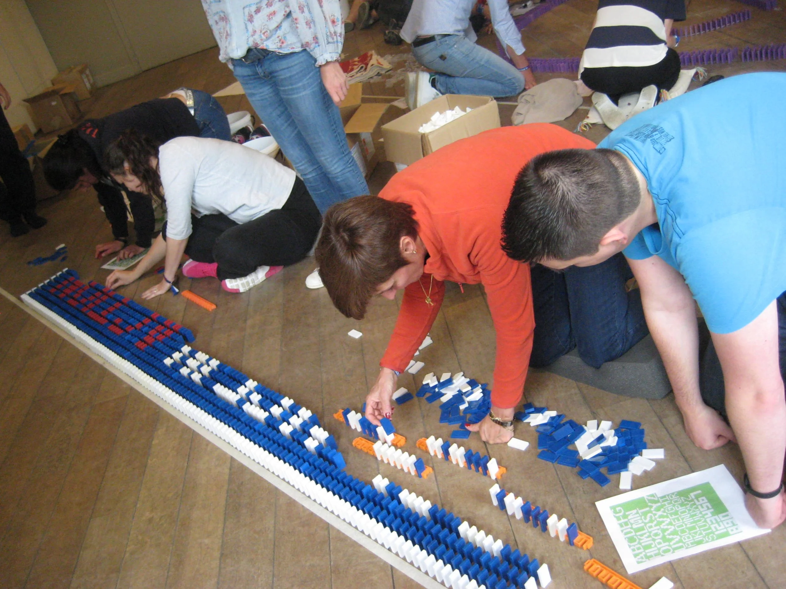 Mensen bouwen een dominolijn met blauwe, witte en oranje dominostenen op een houten vloer.