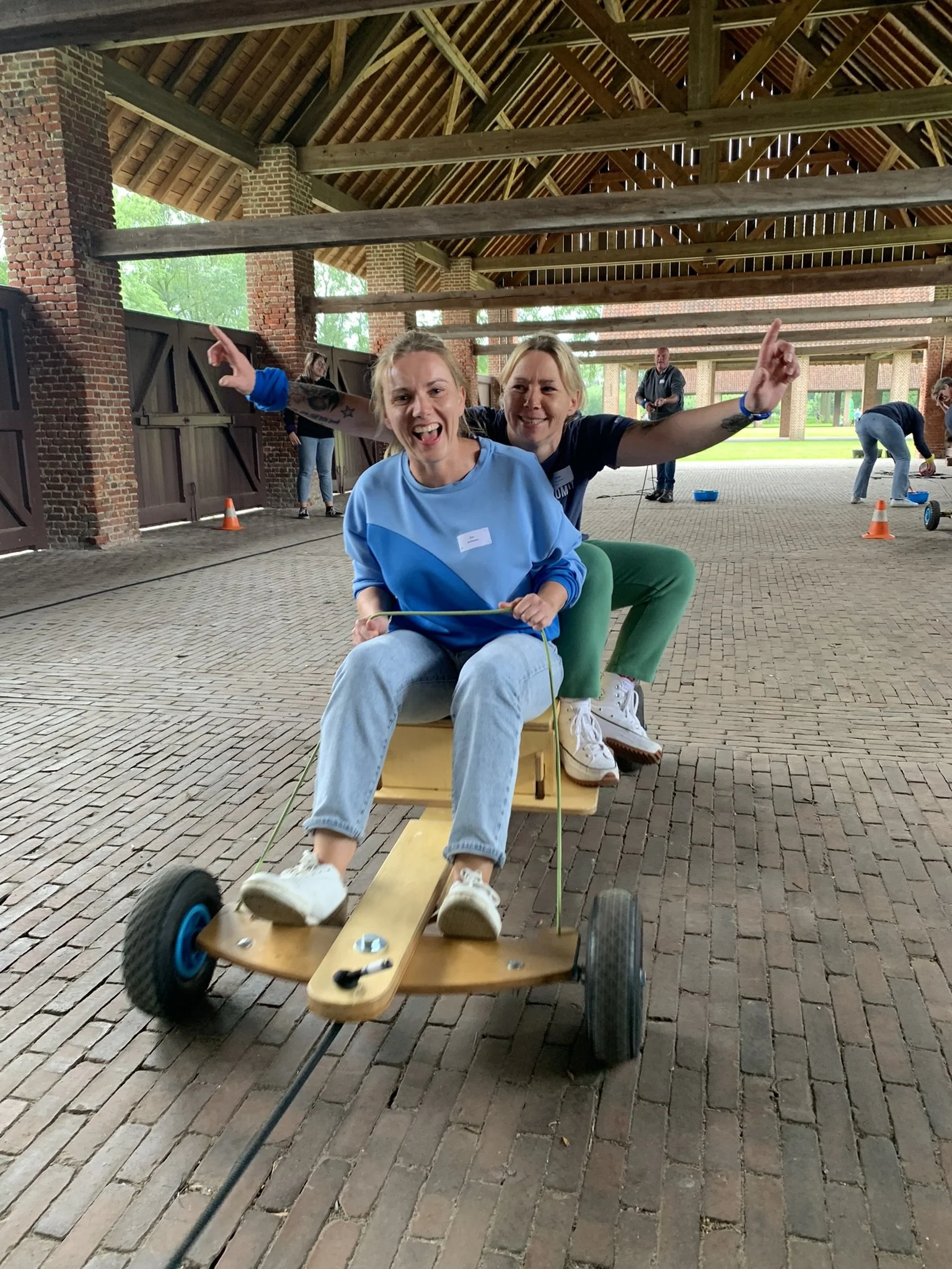 Twee jonge vrouwen genieten van een rit op een houten kar met drie wielen, met een onder een overkapping in een open bouwplaats. Ze lachen vrolijk terwijl een vrouw achter haar hand uitsteekt.