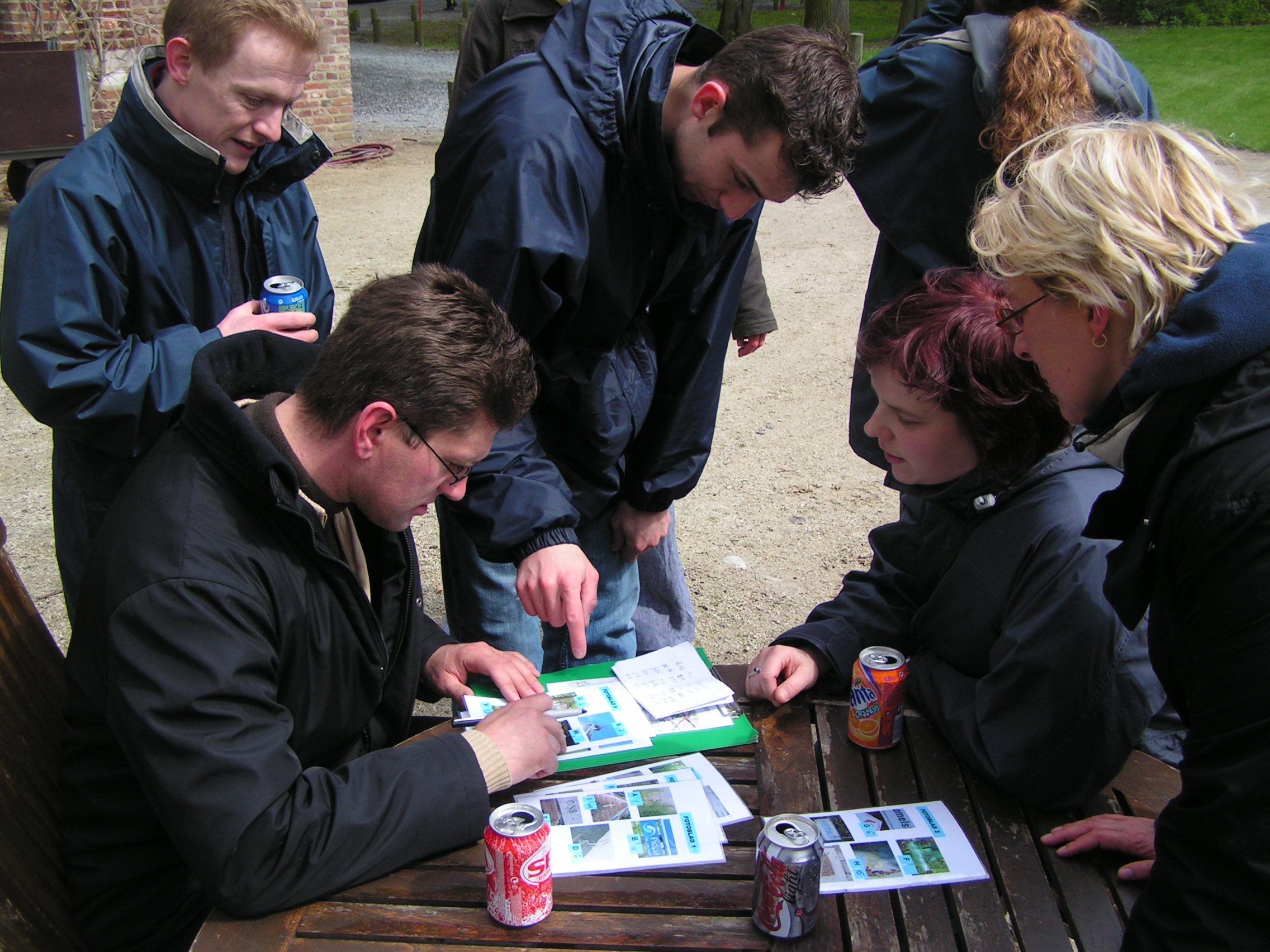 Group of people dressed in coats gathered around a table outside, looking at papers with pictures, some with soda cans.