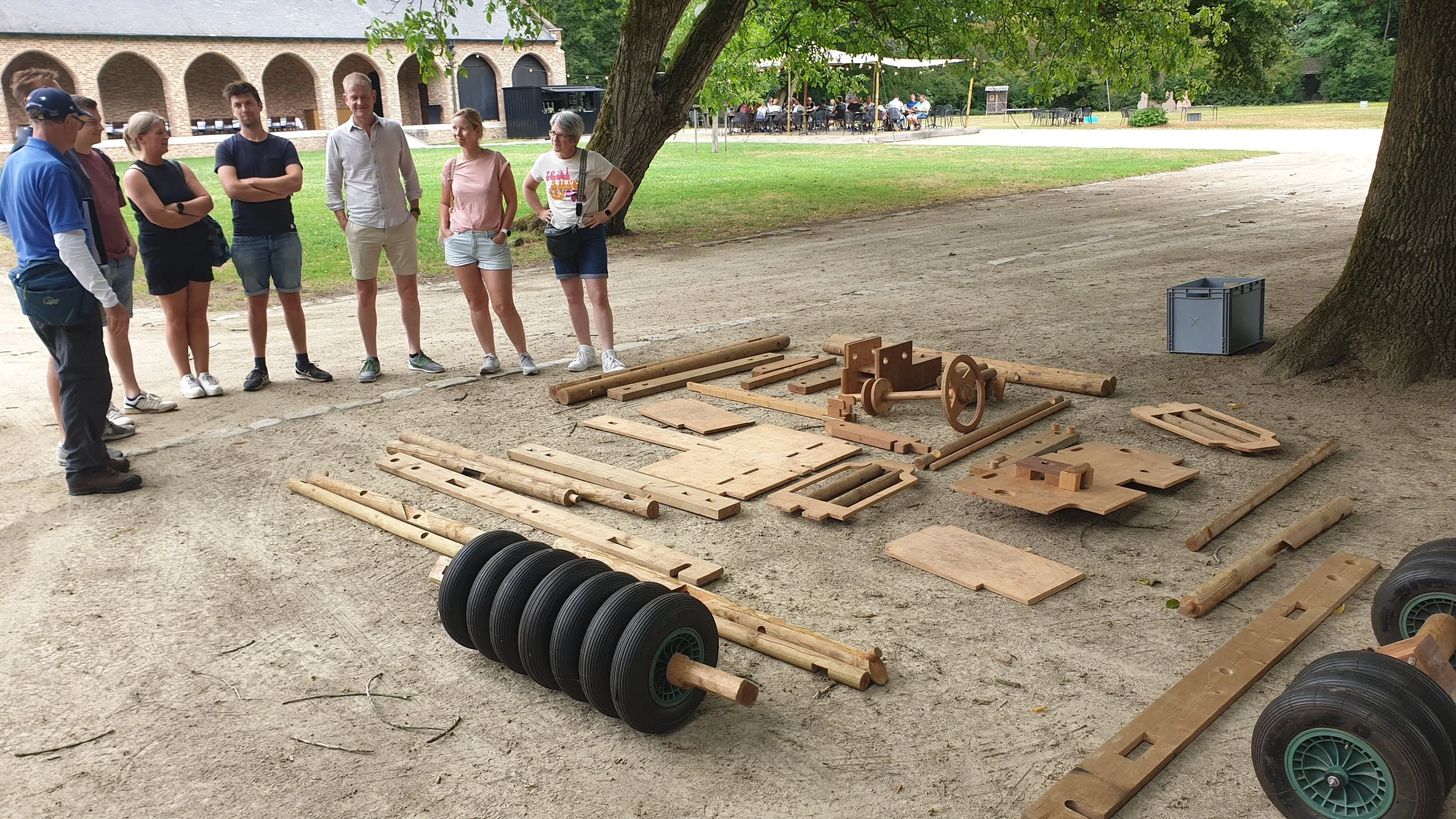 Een groep mensen staat in een cirkel rondom verschillende houten en rubberen bouwmaterialen op de grond in een park, met bomen en een gebouw op de achtergrond.