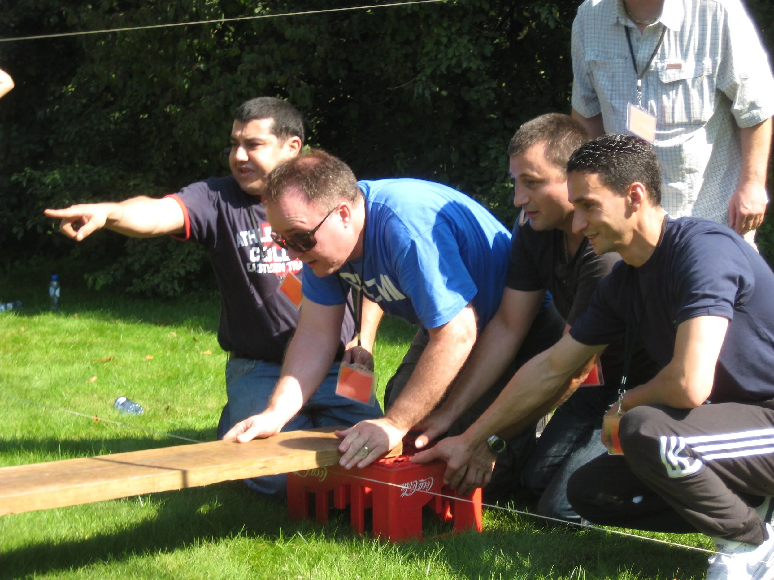 Group of men participating in an outdoor team-building activity, using crates and a plank, on a grassy area.