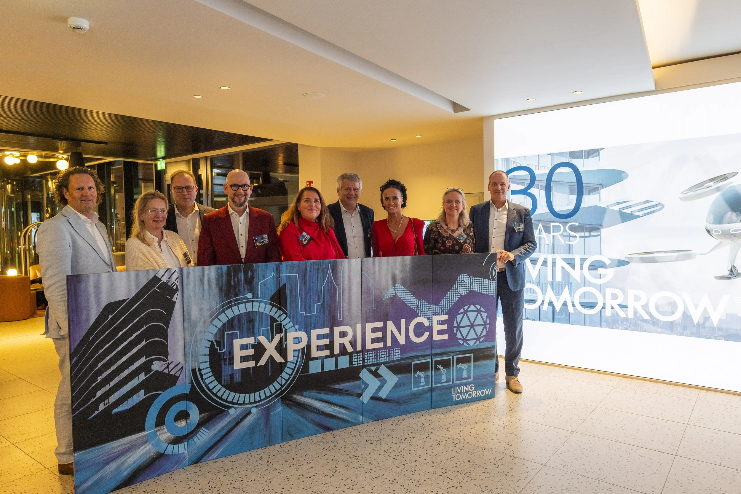 Groep mensen staat achter een blauw decor met het woord 'Experience' en een logo, tijdens een evenement ter gelegenheid van 30 jaar Living Tomorrow in een moderne hal.