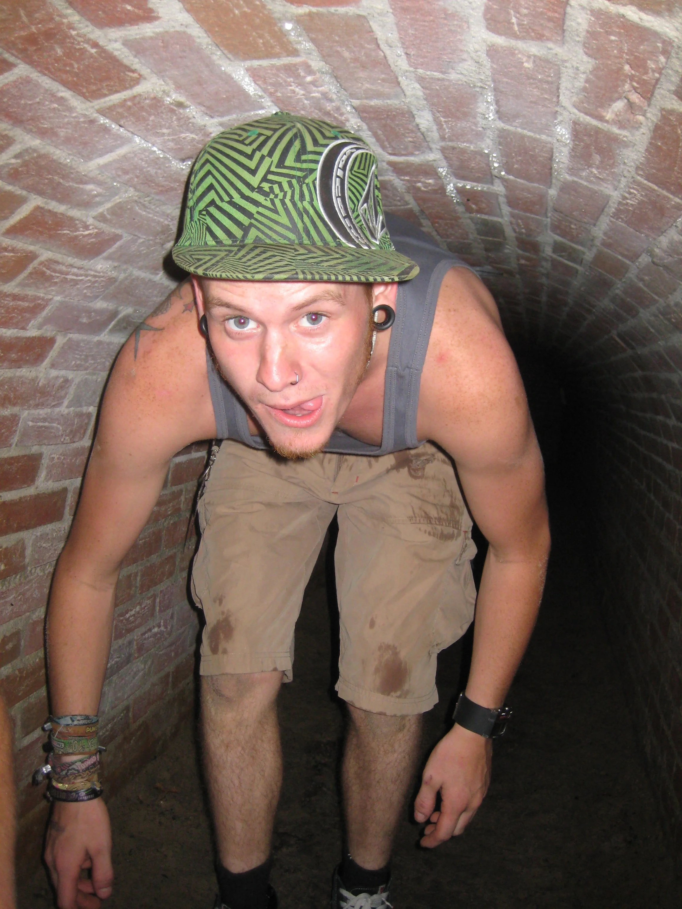 Een man met een groene pet die in een smalle, lage tunnel of ruimte kruipt.