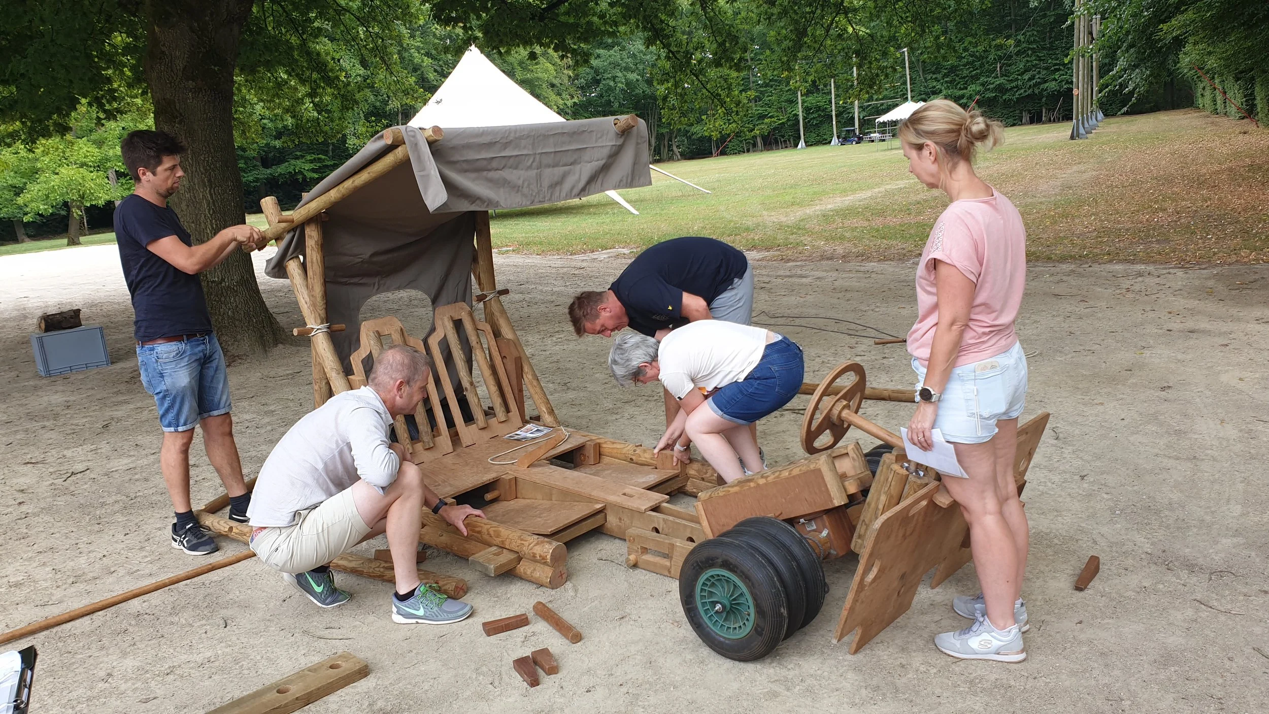 Vijf mensen bouwen samen een houten kar in een park, omringd door bomen en gras, met een grote boom en tenten op de achtergrond.