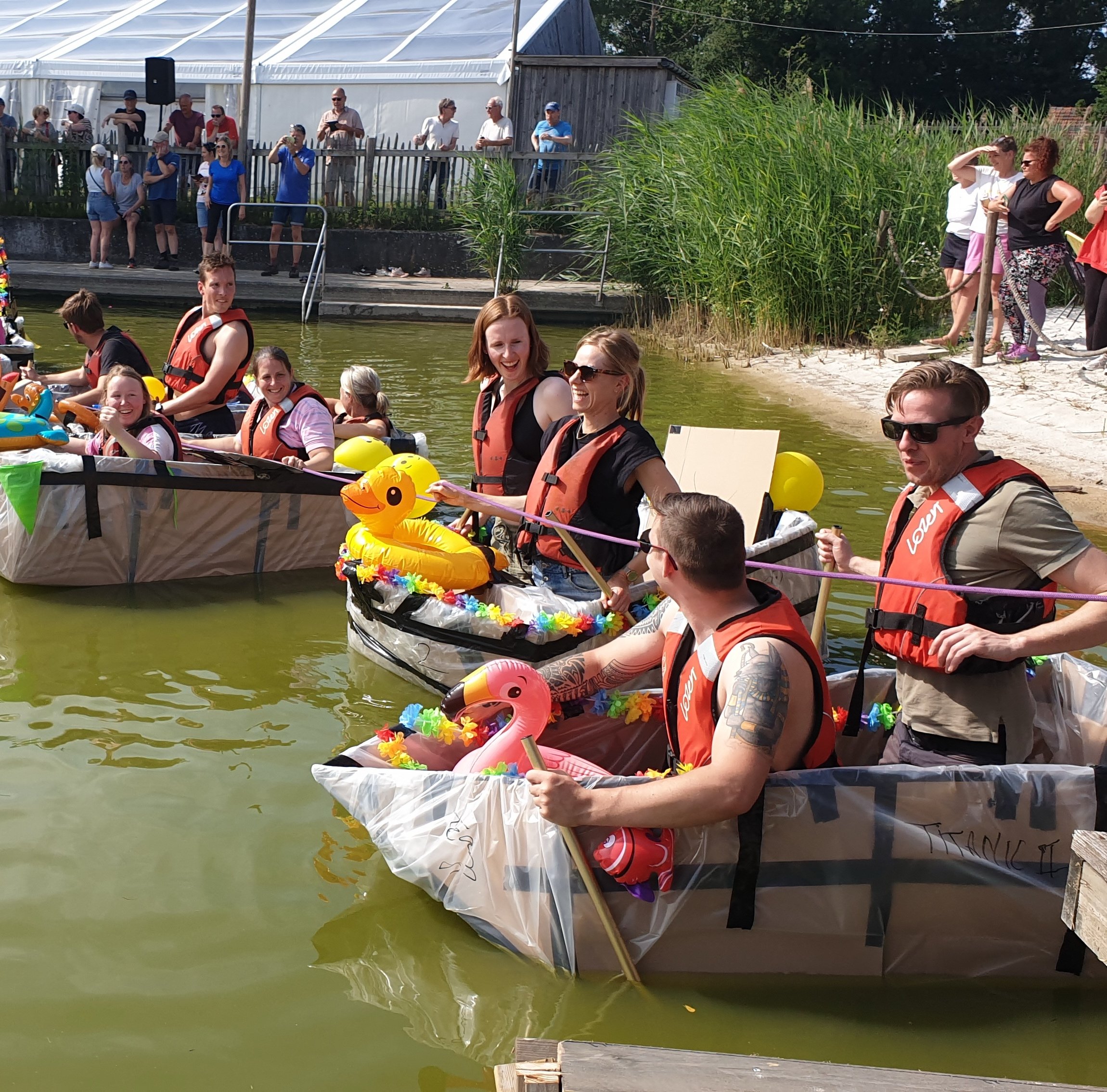 Mensen in zelfgemaakte kartonnen boten varen op een smalle rivier, met groene oevers.