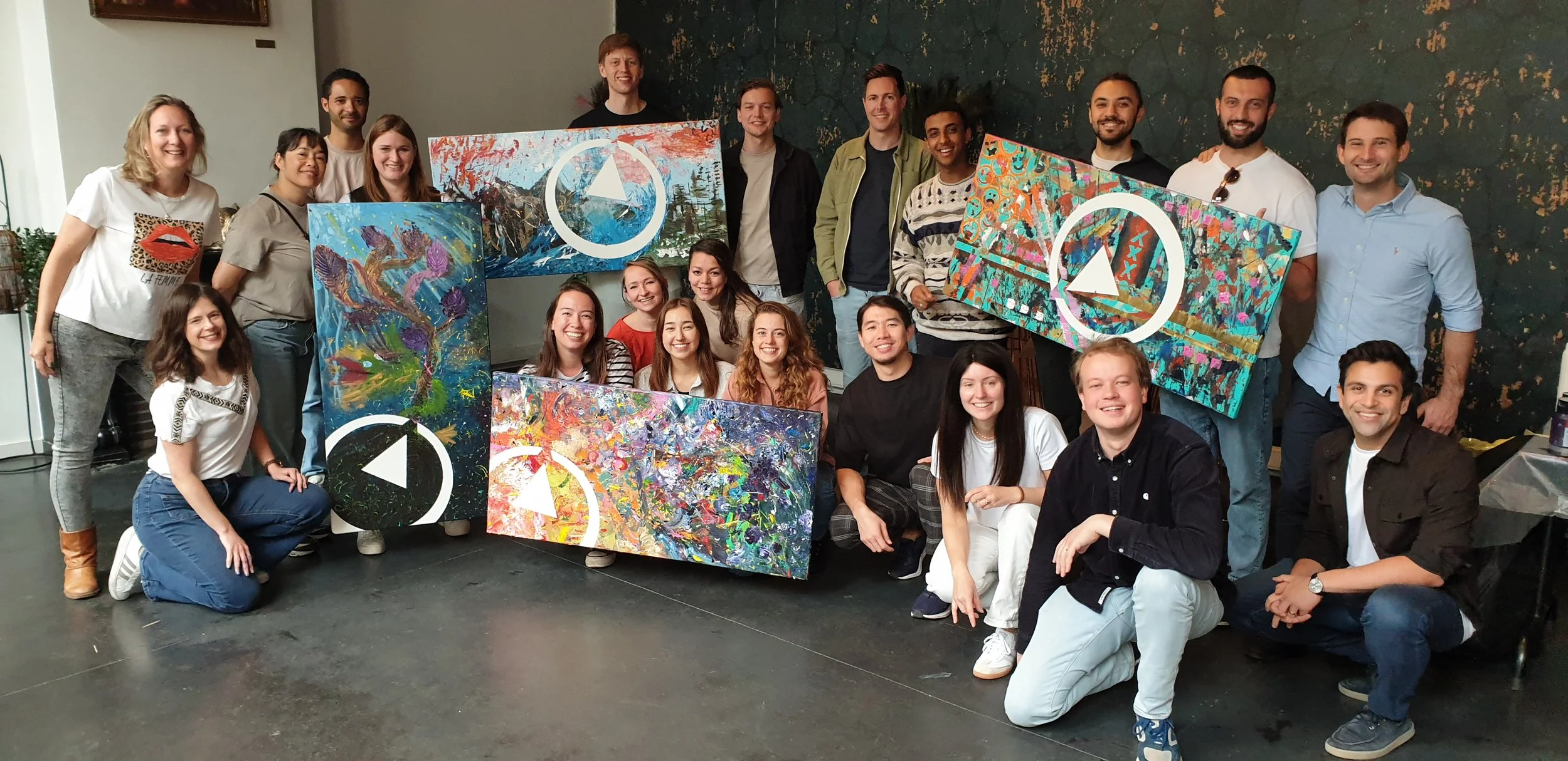 Een groep mensen poseert samen binnenshuis met kleurrijke schilderijen. Ze zijn allemaal glimlachend en het lijkt op een creatieve of sociale bijeenkomst. De schilderijen hebben verschillende abstracte ontwerpen met felle kleuren.