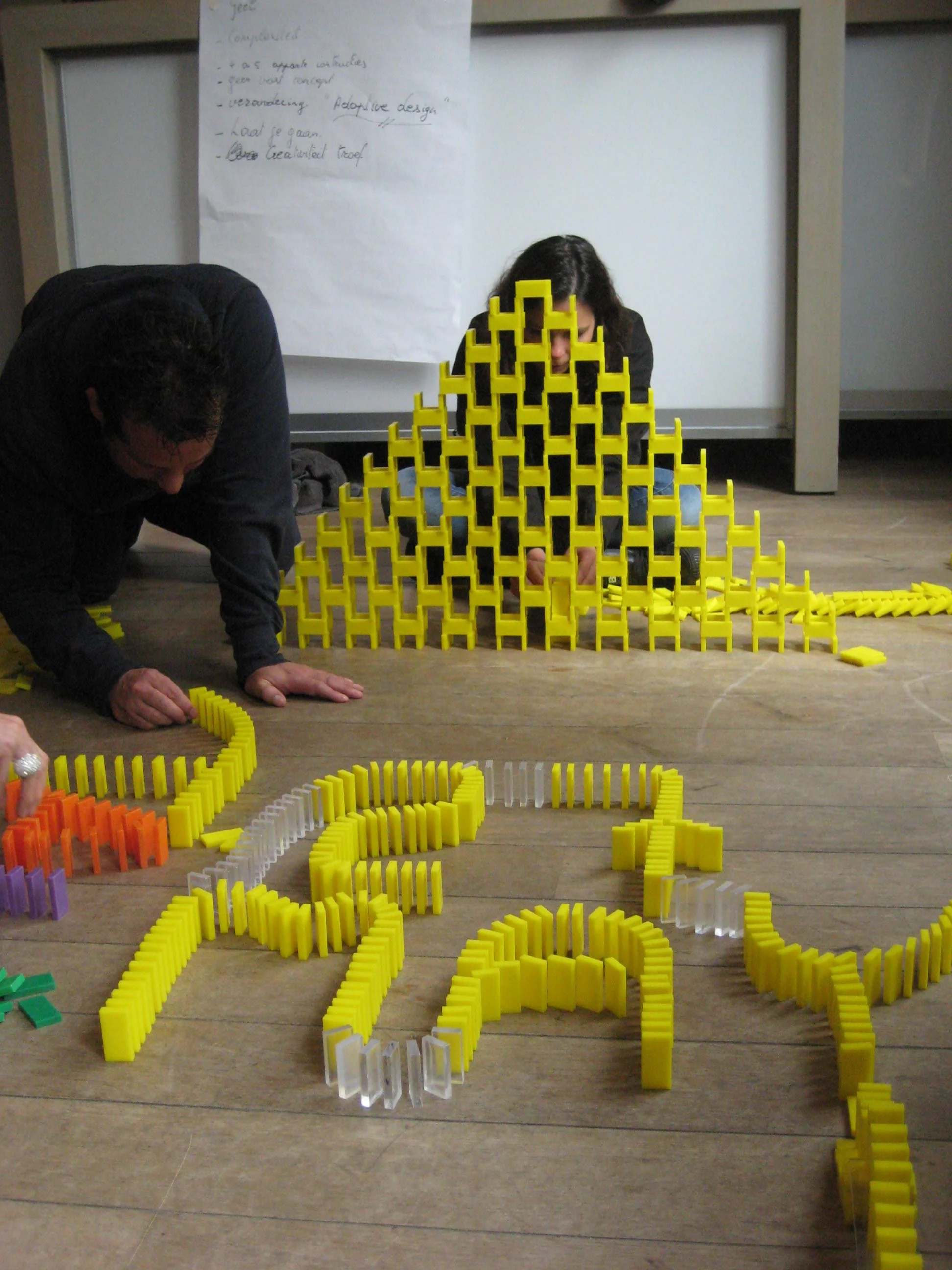 Twee mensen bouwen een toren en woorden met gele dominostenen. Sommige stenen zijn ook oranje of paars. Er staat een groot geschreven vel papier op de achtergrond.