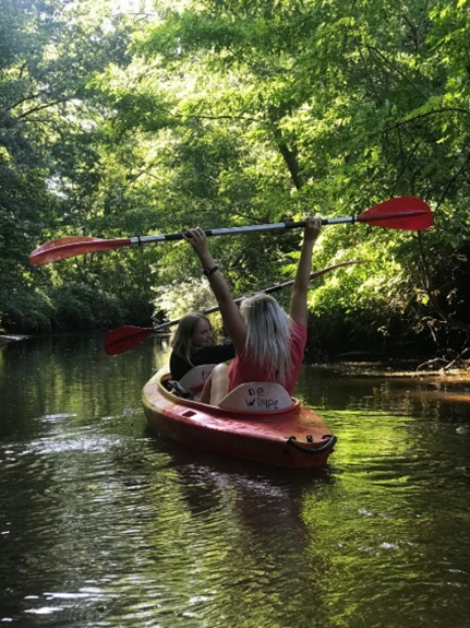Twee kinderen in een rode kajak op een rustige rivier, omringd door groene bomen, met de ene kind die een lange peddel omhoog houdt.