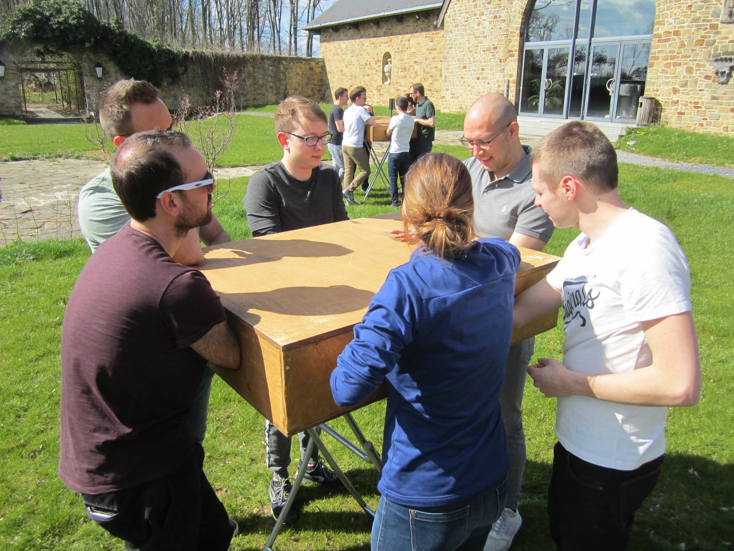 Groep mensen die rond een tafel buiten staan, bezig met een activiteit. Ze bevinden zich op een grasveld voor een stenen gebouw met grote ramen. Het is zonnig weer.