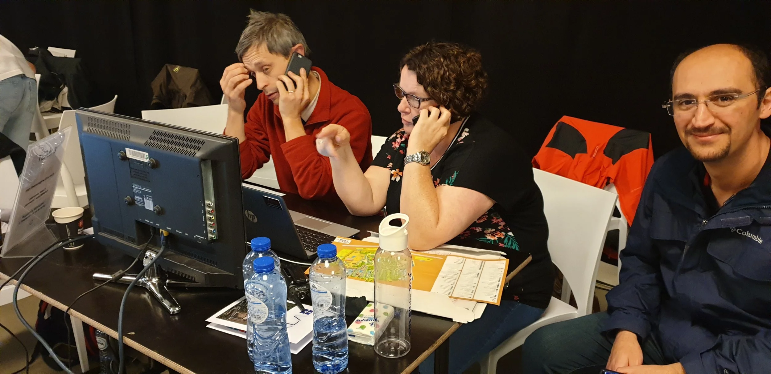 Drie mensen zitten aan een tafel met computers en documenten, allemaal met telefoons in de hand, in een vergaderruimte.